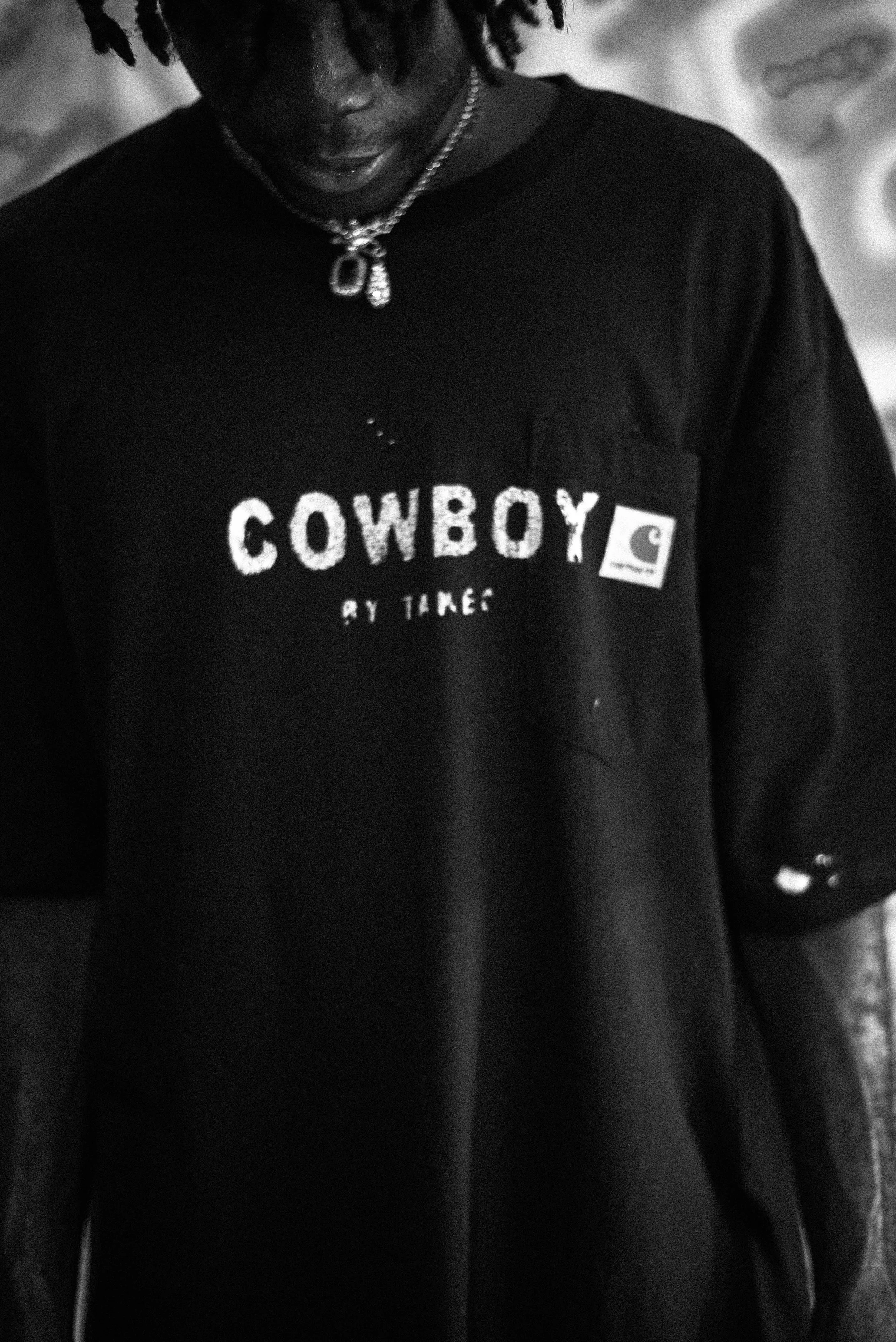 A person wearing a black t-shirt with the word 'COWBOY' printed on it, a Carhartt logo patch, and a silver necklace, with dreadlocks visible.
