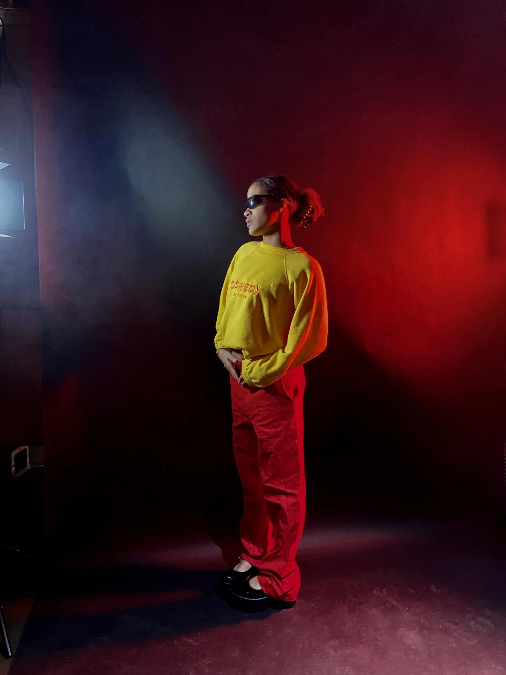 A woman wearing sunglasses, a yellow sweatshirt, and red pants standing against a red and dark background.