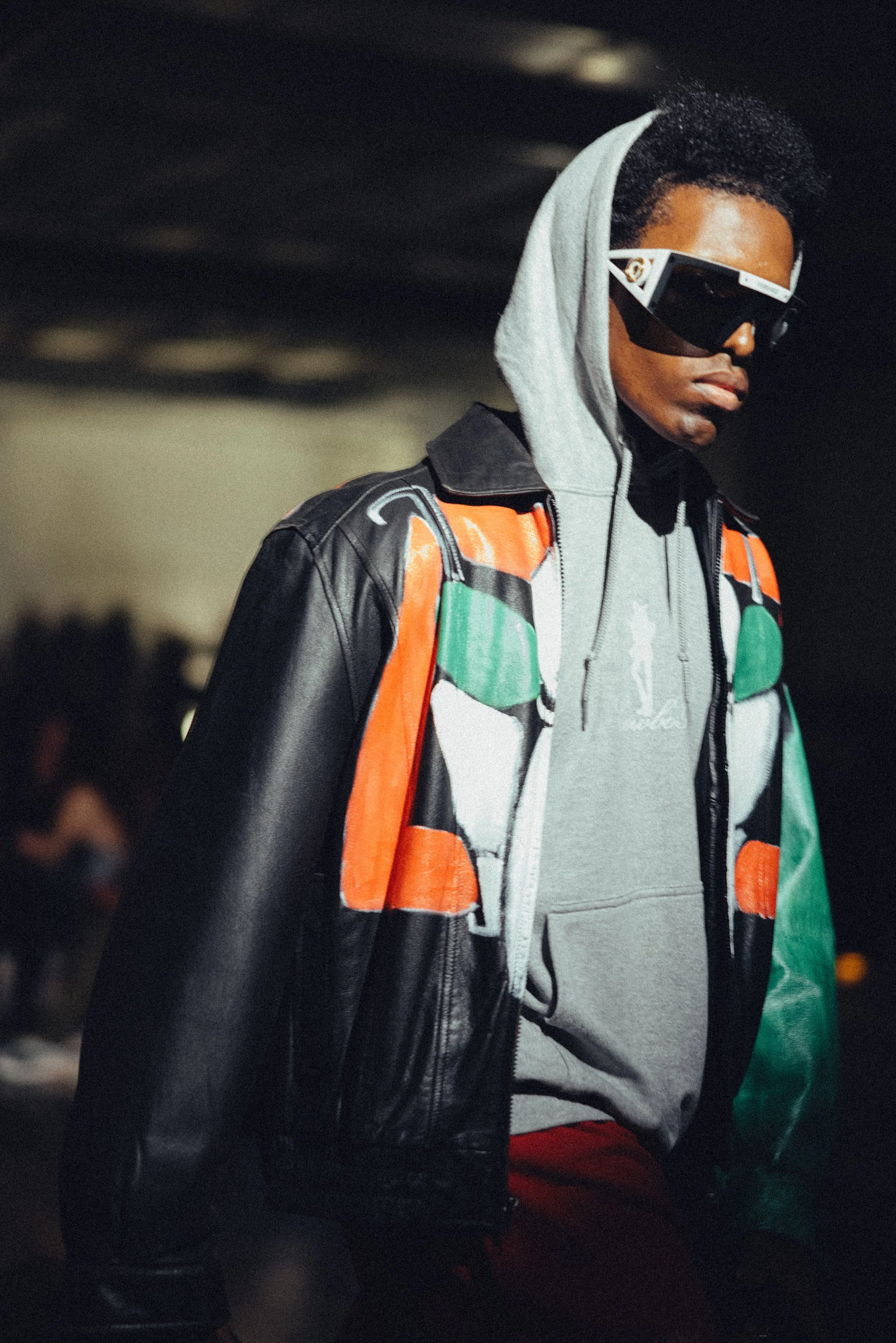 A person wearing large black sunglasses, a gray hoodie, a multicolored jacket, and red pants, standing outdoors.