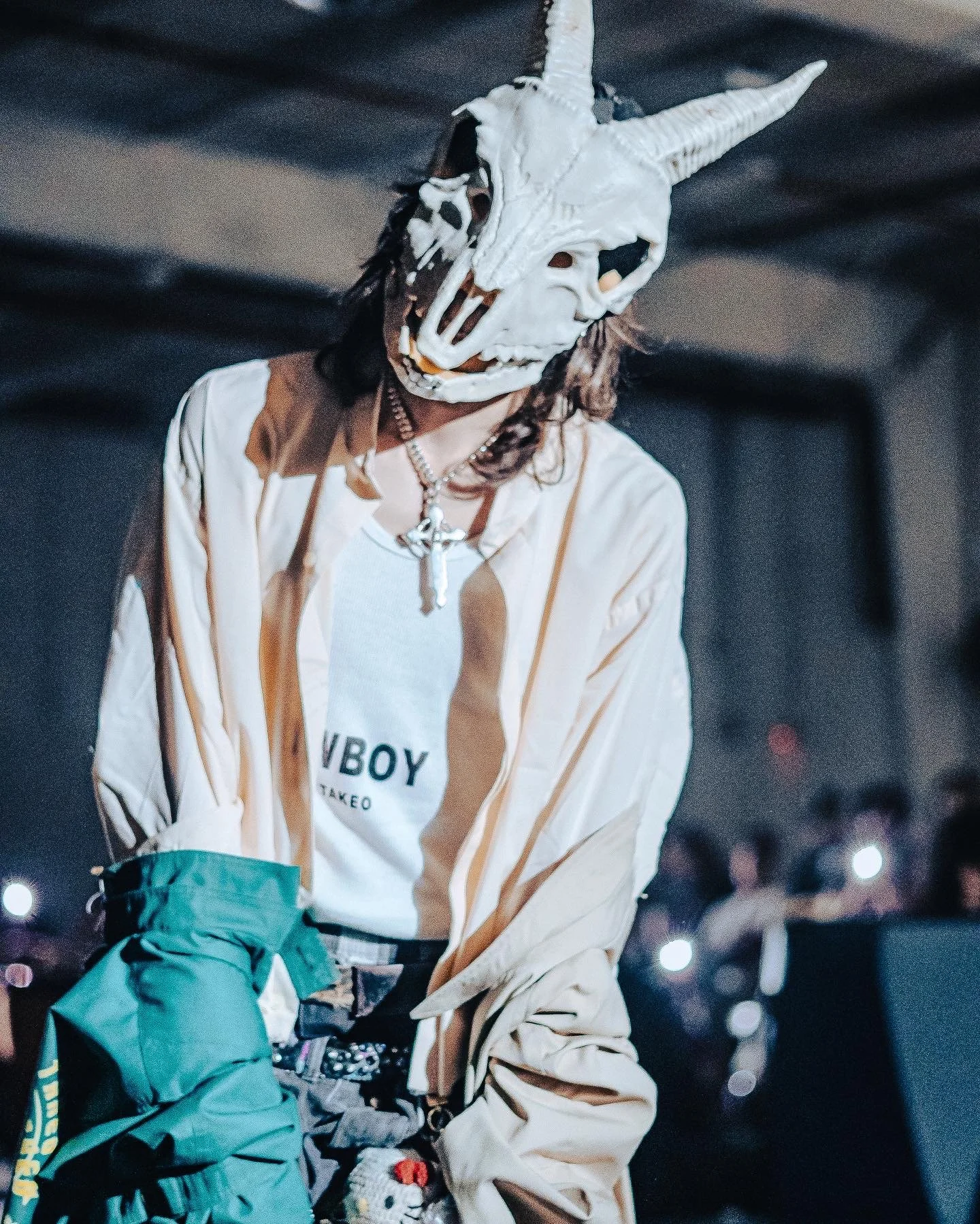 Person wearing a goat skull mask with horns, a white T-shirt, beige jacket, and several necklaces, in a dark indoor setting with other people in the background.