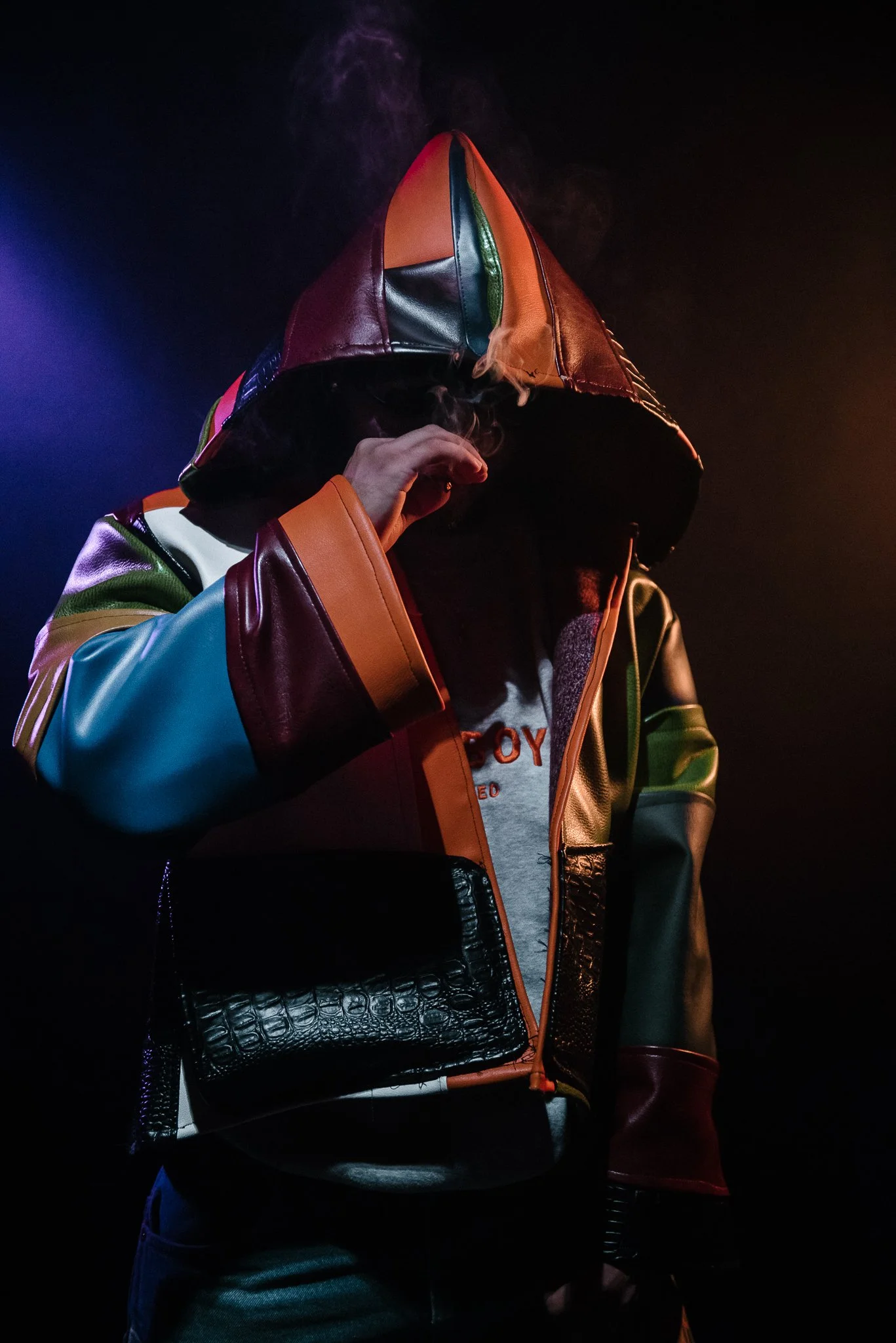 Person wearing a multicolored leather jacket and hood, partially covering face, against a dark background with colorful lighting.
