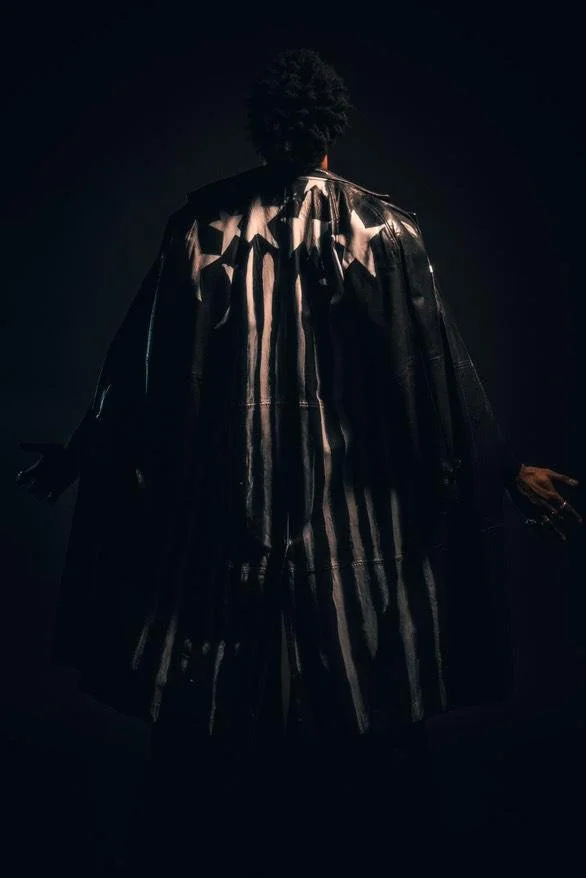Back view of a person wearing a black leather coat with white stars and black and white vertical stripes, standing in a dark setting.