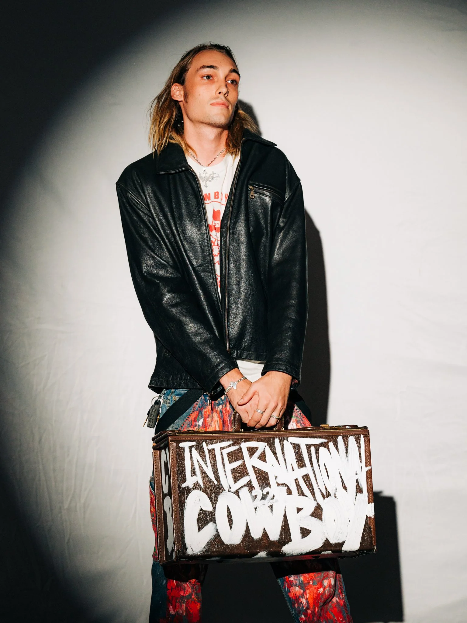 A man with long hair and a leather jacket, holding a suitcase with the words 'International Cowboy' painted on it in white letters.