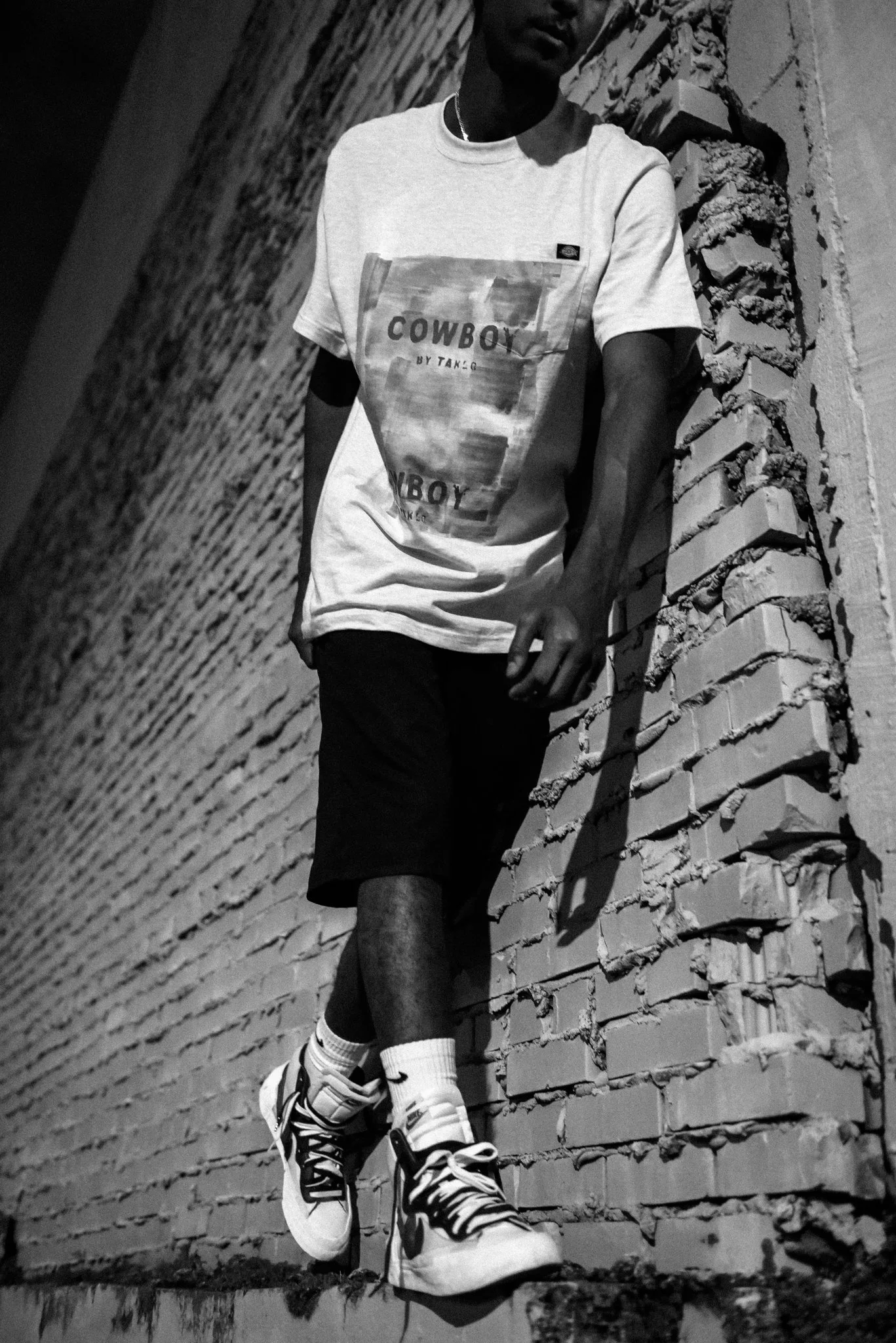A young man wearing a graphic t-shirt, shorts, and sneakers leans against a brick wall with graffiti or peeling paint, in a black and white photo taken at an angle.