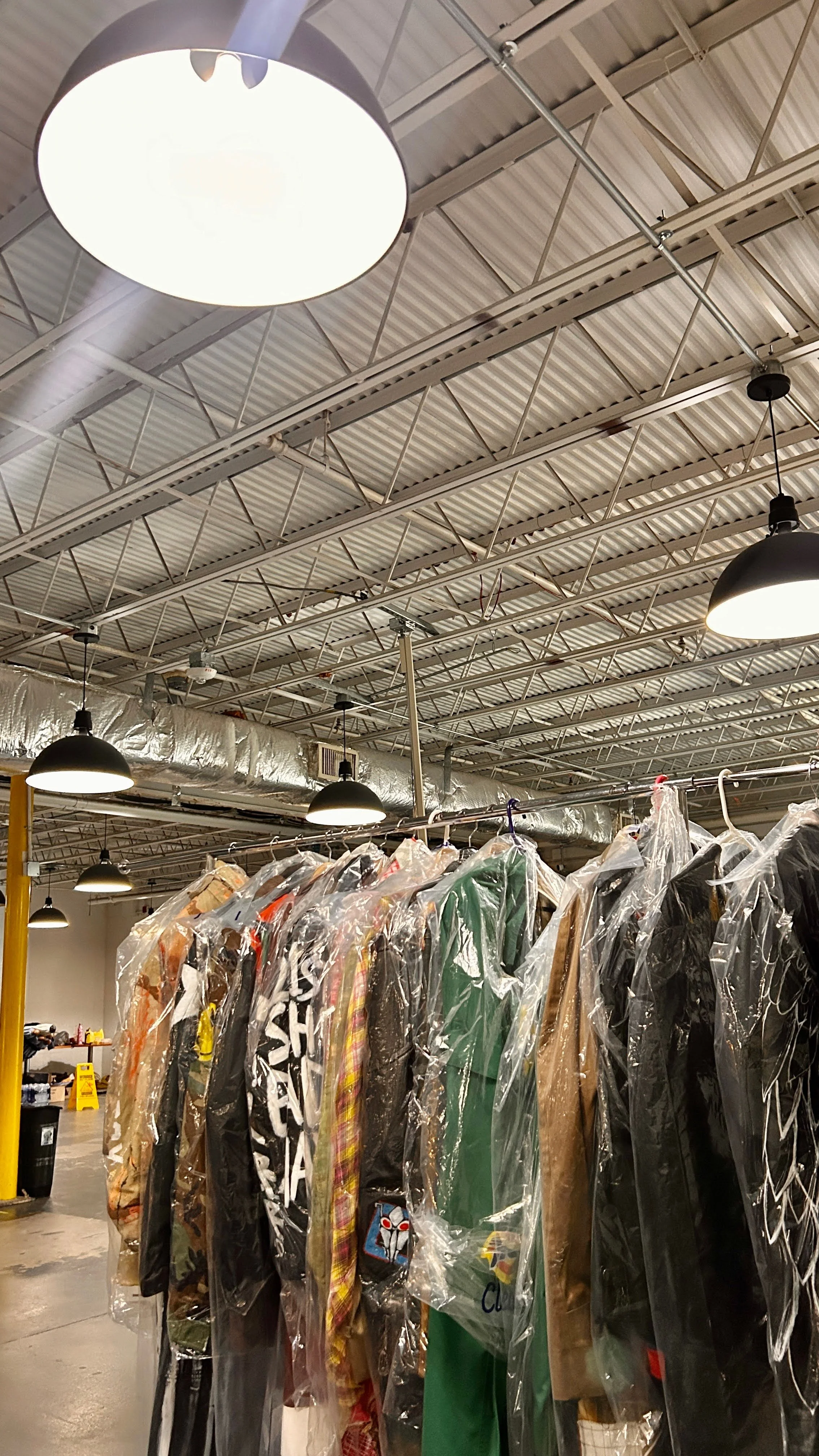 Clothing items hanging on a rack, covered used plastic at a thrift store or clothing warehouse, with industrial ceiling lights and exposed ventilation ductwork overhead.