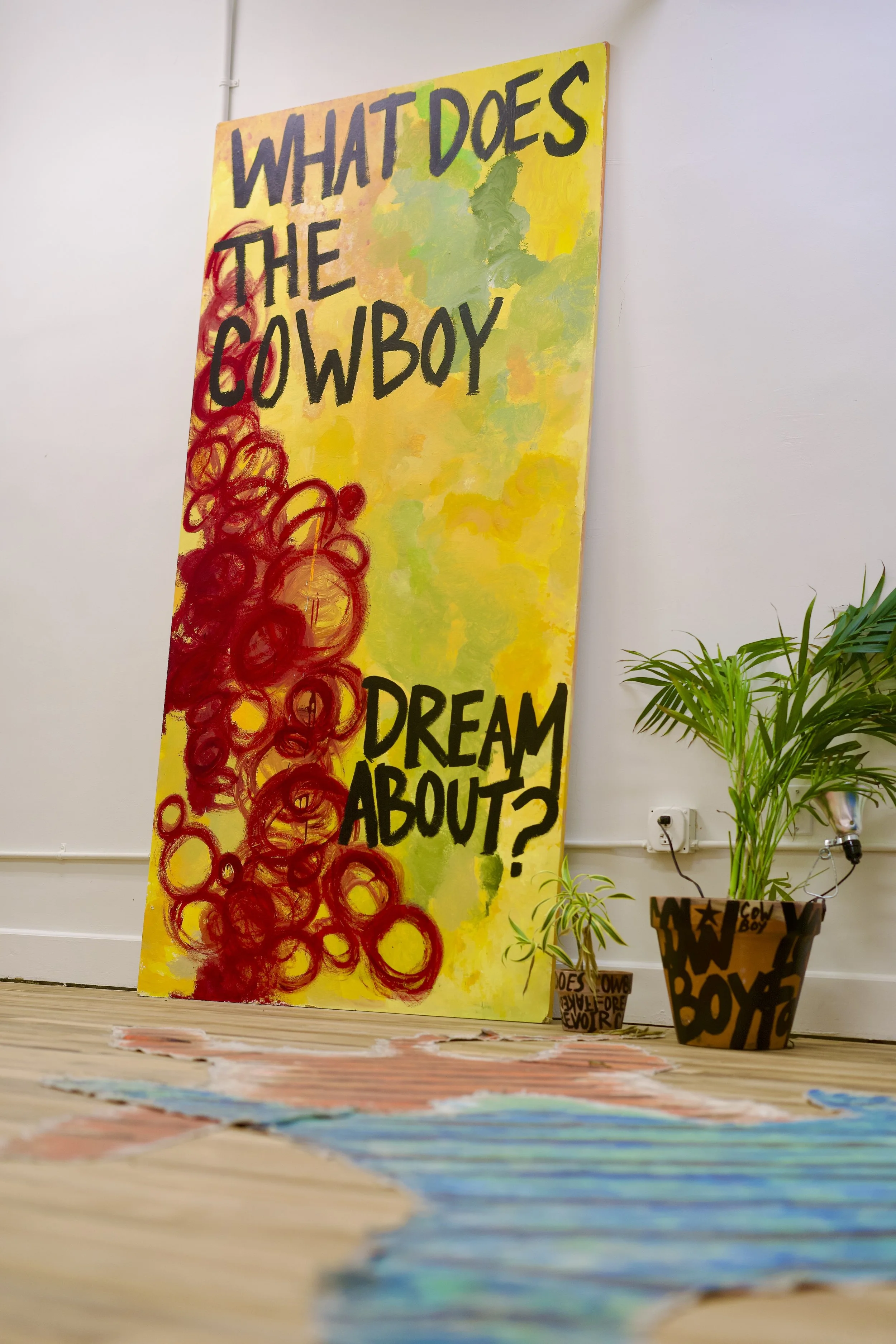 A colorful acrylic painting with the words "What does the cowboy dream about?" in bold black letters, with a background of green, yellow, and red swirls, leaning against a white wall near potted plants.