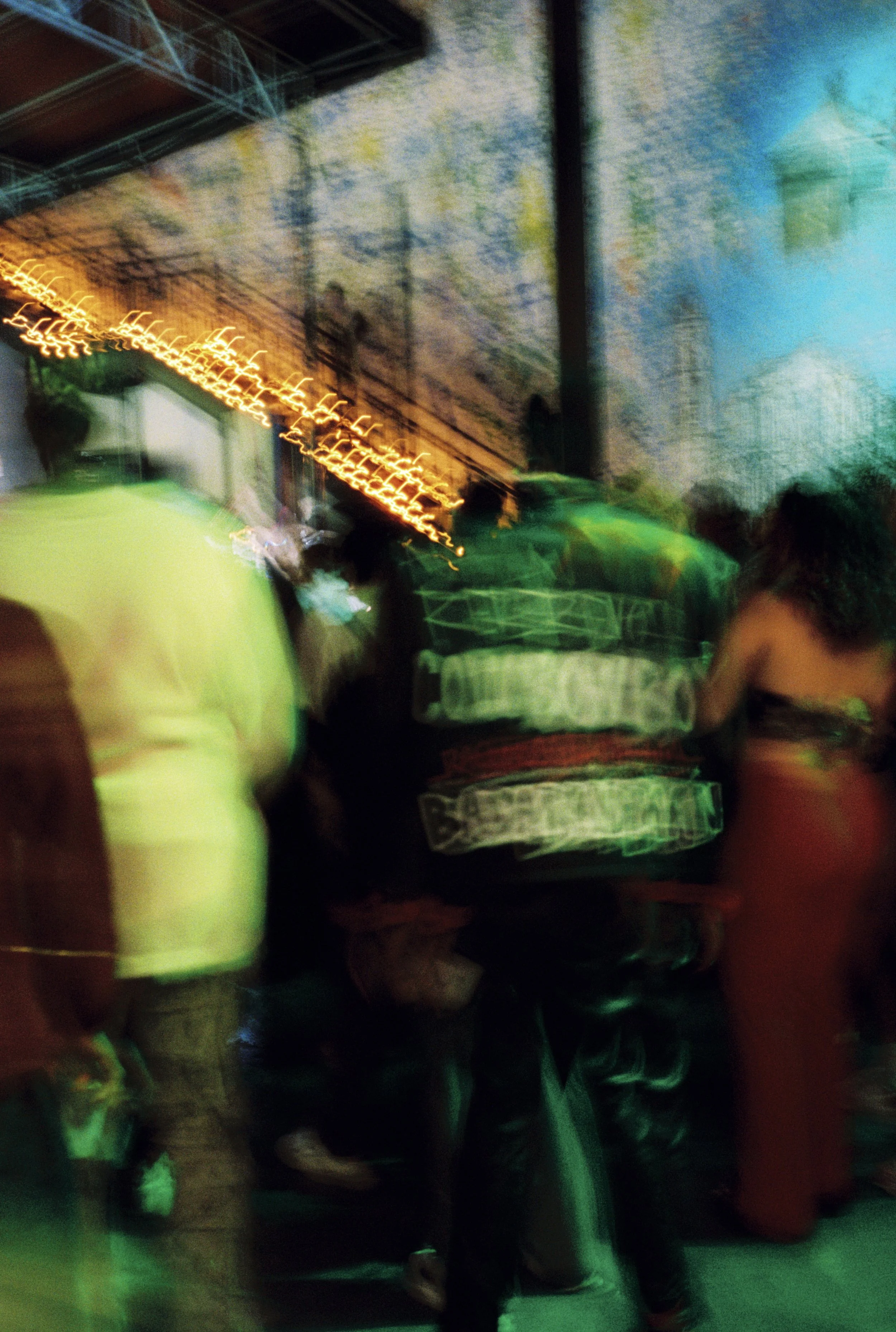 Blurry photo of people at a crowded indoor event with colorful lighting and a staircase in the background.