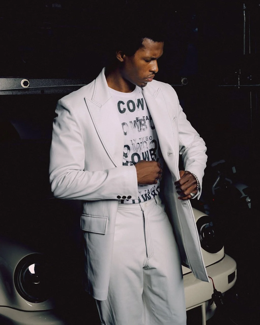 A man in a white suit and T-shirt stands with hands adjusting his coat, positioned in front of a small white car in a dark setting.