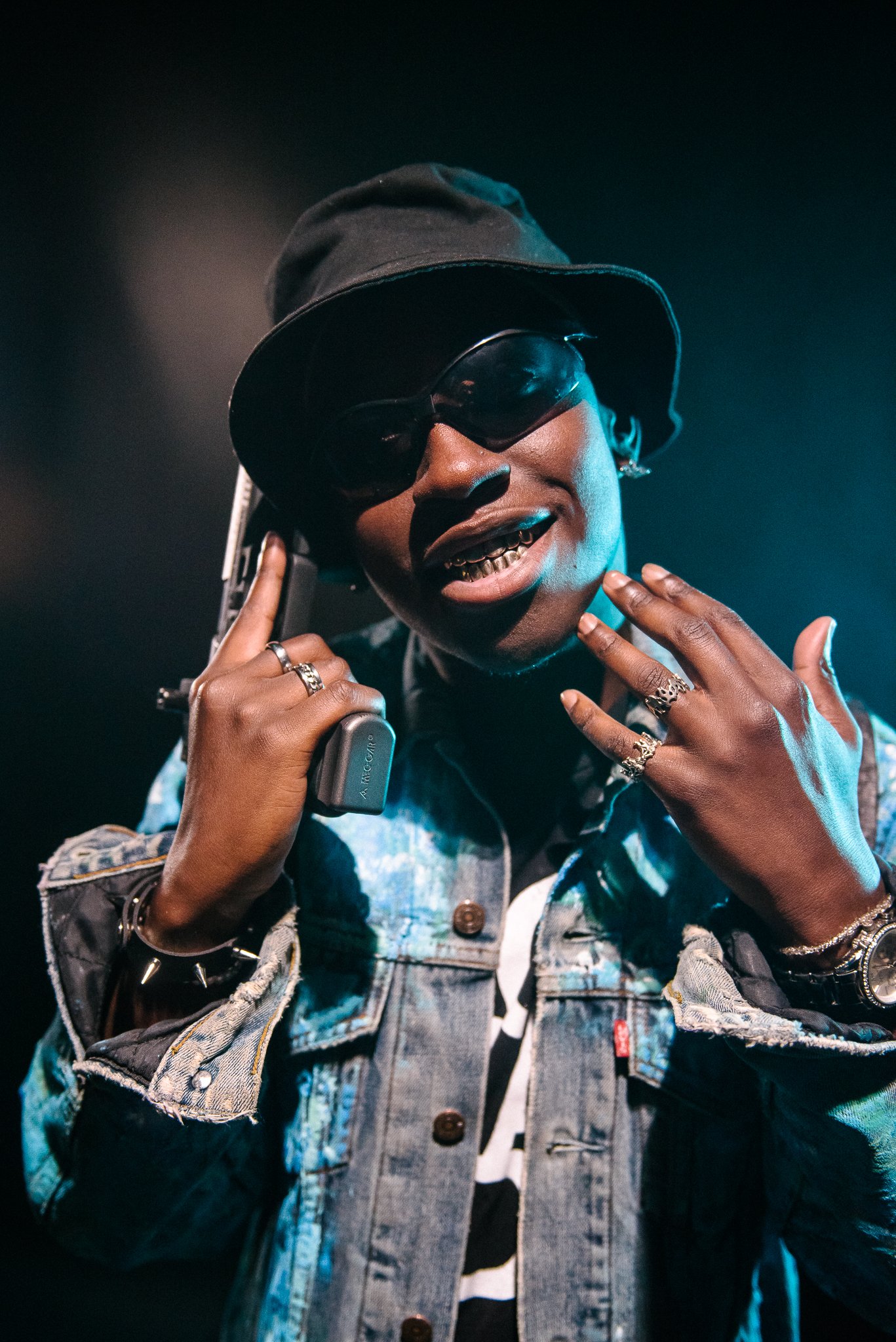 A person wearing a black hat and sunglasses, holding a vintage phone to their ear, smiling with visible braces, adorned with rings, bracelets, and watch, dressed in a denim jacket over a striped shirt, standing against a dark background.