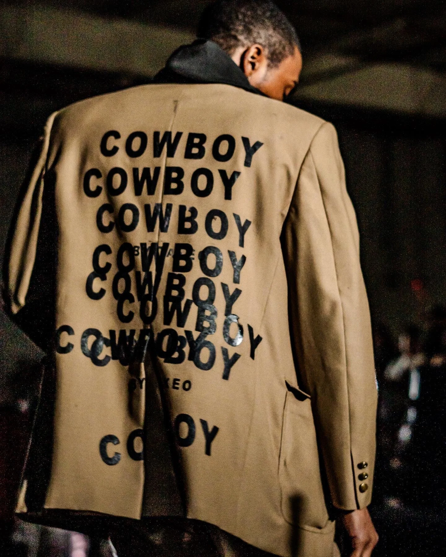 A man seen from behind wearing a beige blazer covered in black 'COWBOY' text in various sizes and orientations.