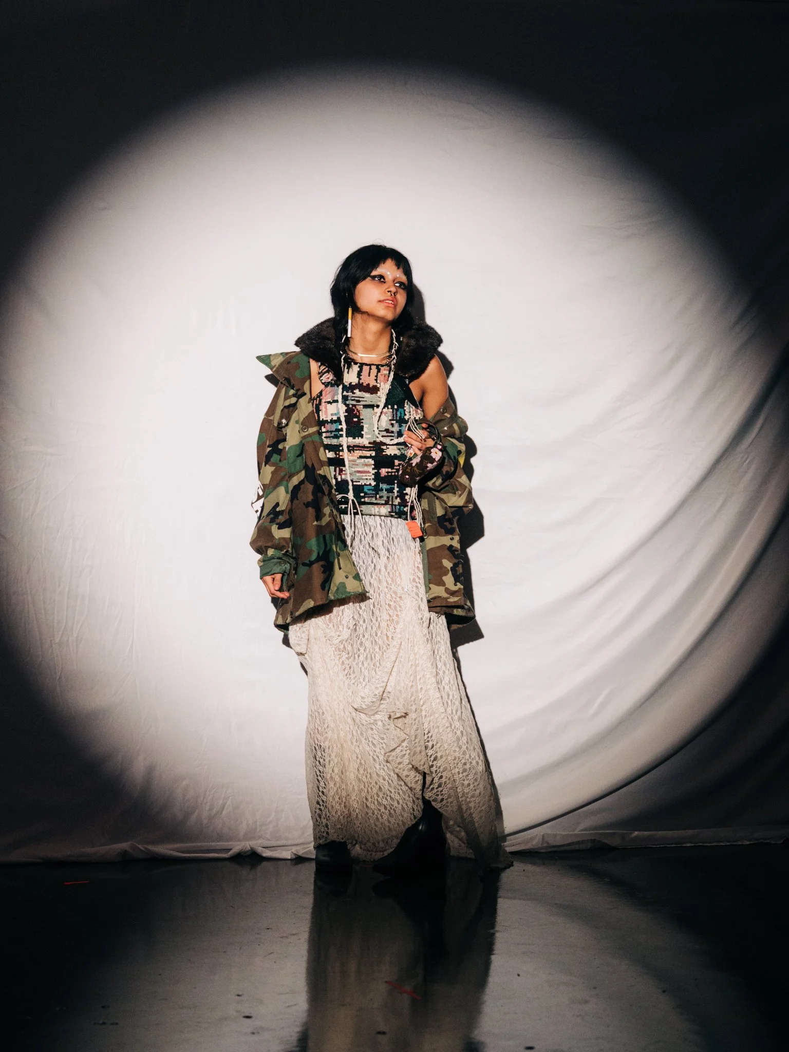 Woman standing in front of a white backdrop, viewed through a circular opening, wearing a lace maxi skirt, a patterned top, and a camouflage jacket.