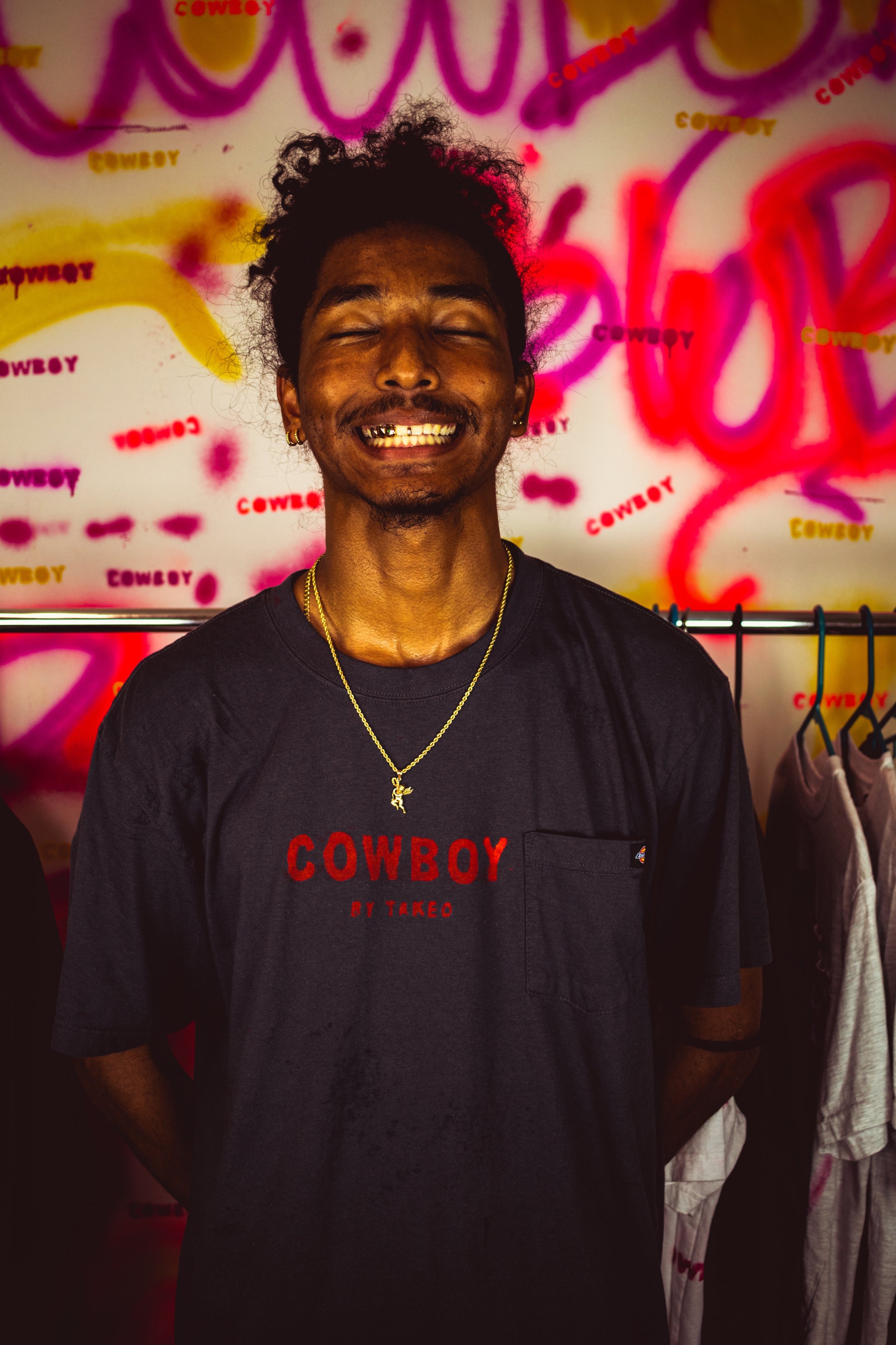 A man with curly hair, gold grillz, and a gold necklace smiling with eyes closed, standing in front of a wall with colorful graffiti that says 'COWBOY' repeatedly.