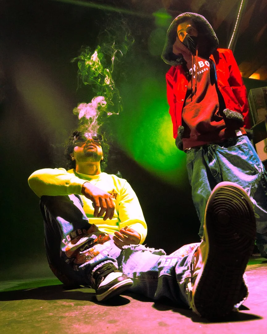 Two young men with hoodies and jackets in a smoky atmosphere illuminated by green and purple lights, one sitting on the floor smoking, the other standing nearby.