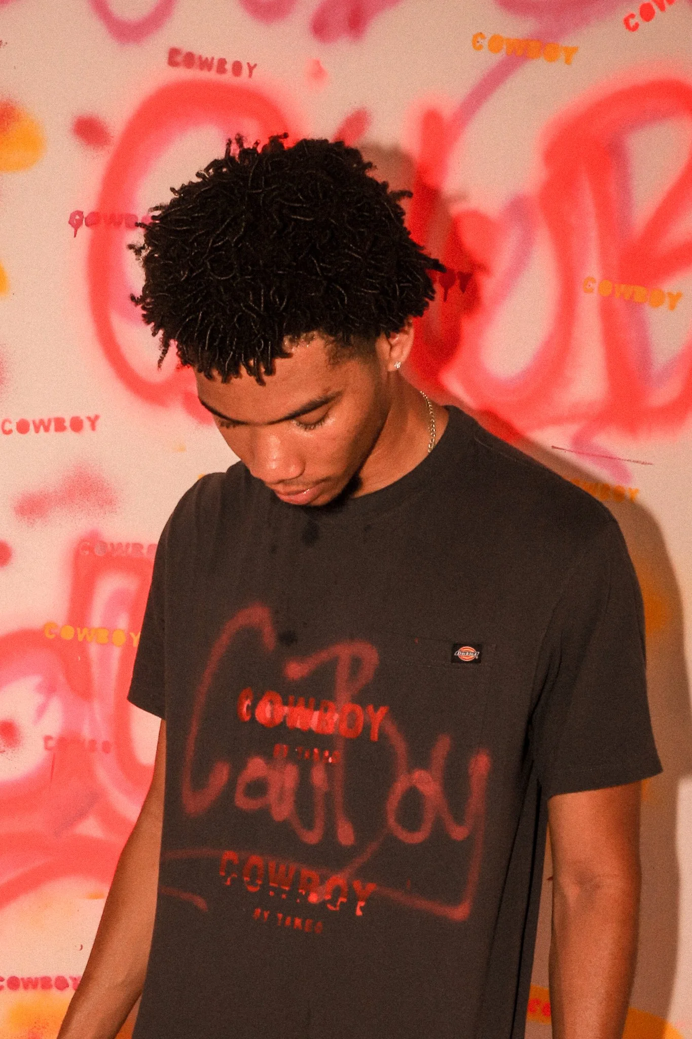 A young man with curly black hair looking down, wearing a black T-shirt with red and white graphics and a small logo, standing in front of a red and yellow graffiti-style background with the words "COWBOY" and other text.