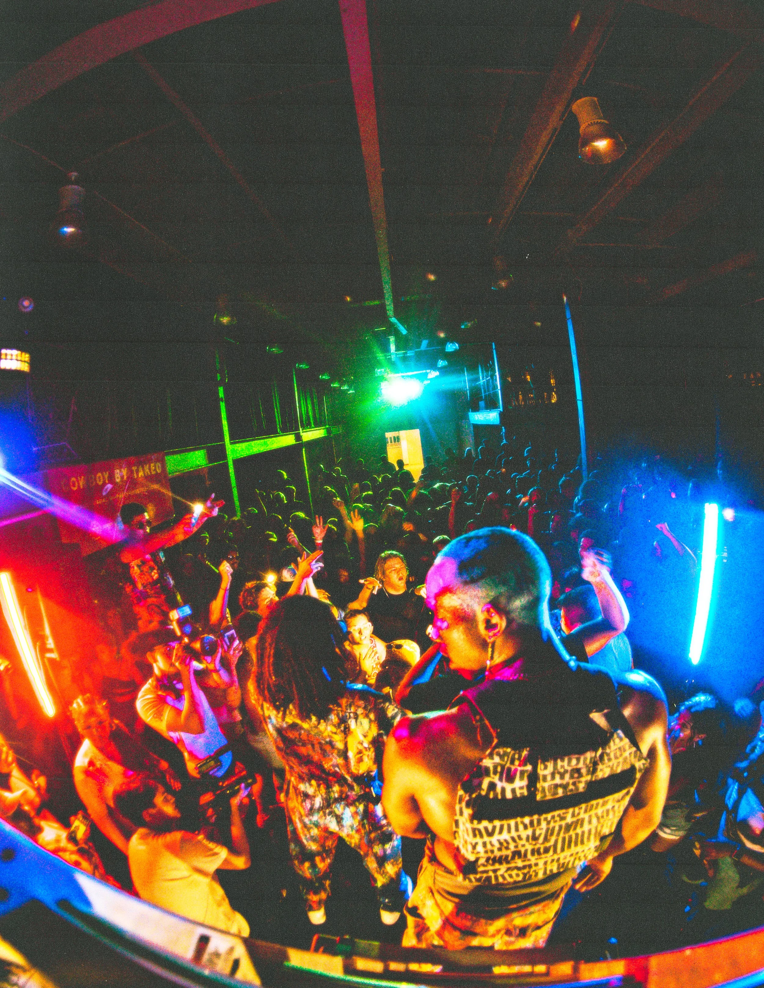 Crowd of people dancing in a nightclub with colorful lights and a DJ booth.