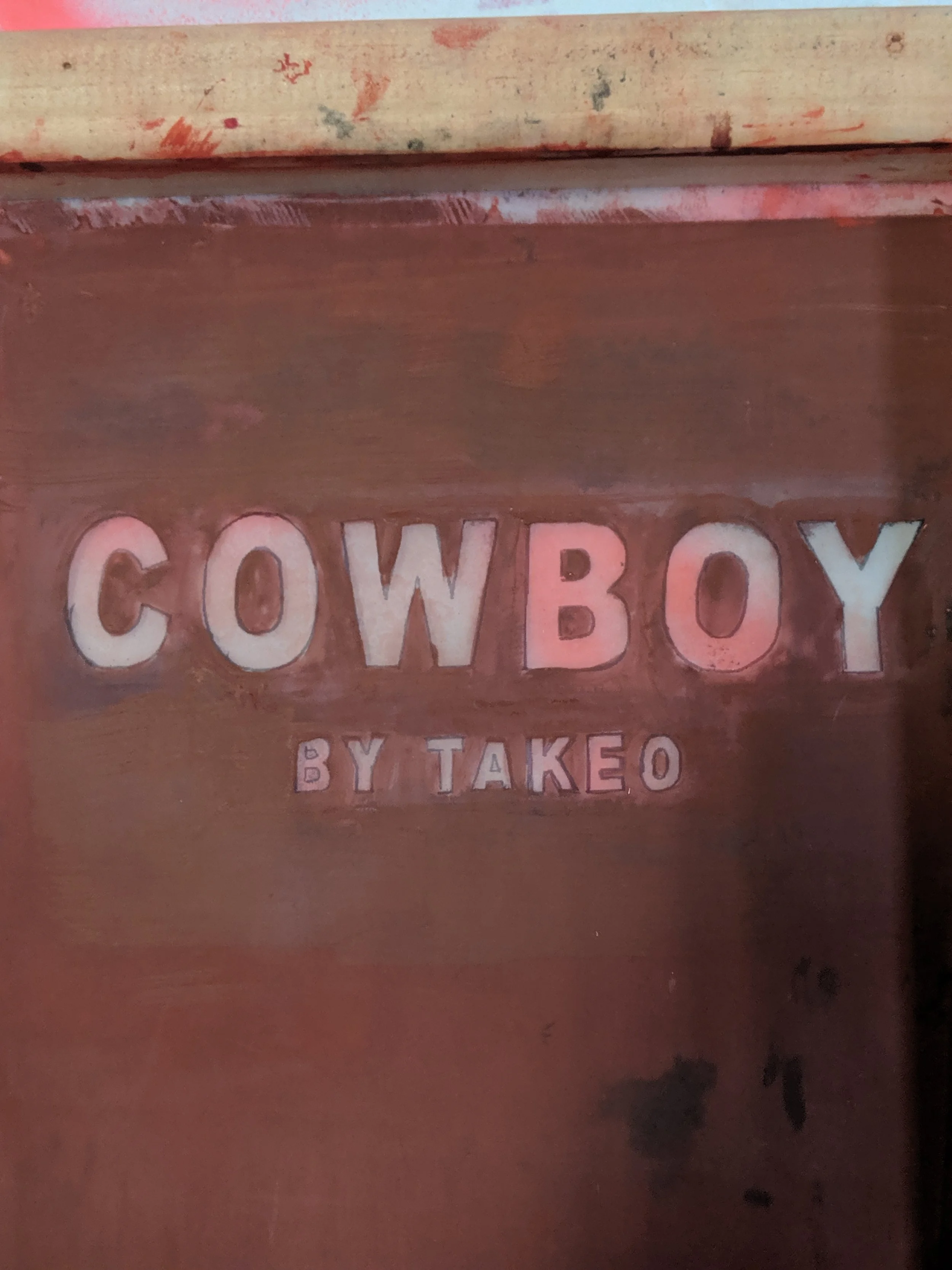 Rustic wooden sign that reads "Cowboy" with smaller letters underneath that say "by Takeo".