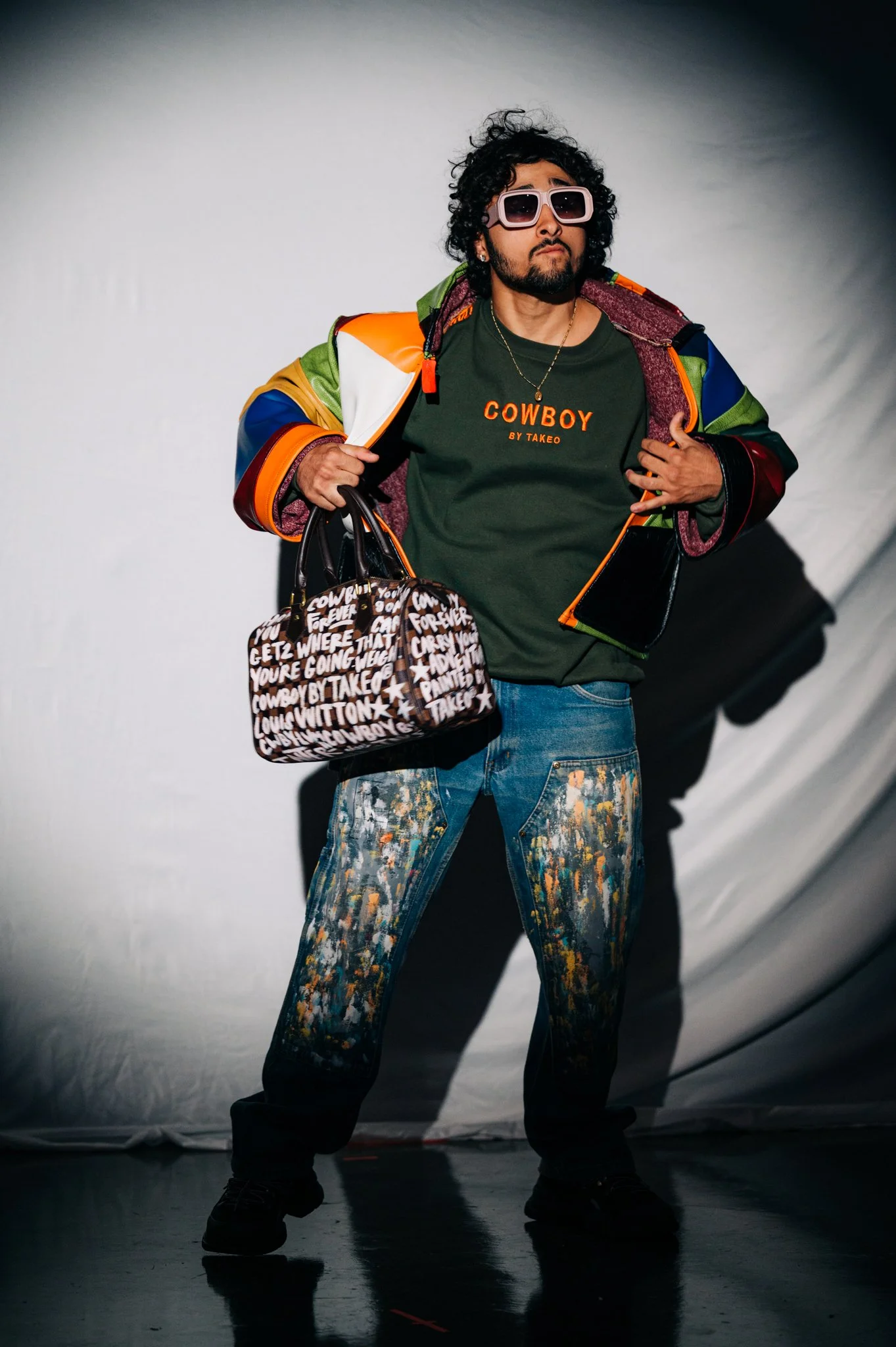 Man with curly hair and sunglasses posing with a confident attitude, wearing a colorful jacket, a green T-shirt with 'COWBOY BY TAKEO' printed on it, paint-splattered jeans, holding a patterned designer bag, standing against a plain background.