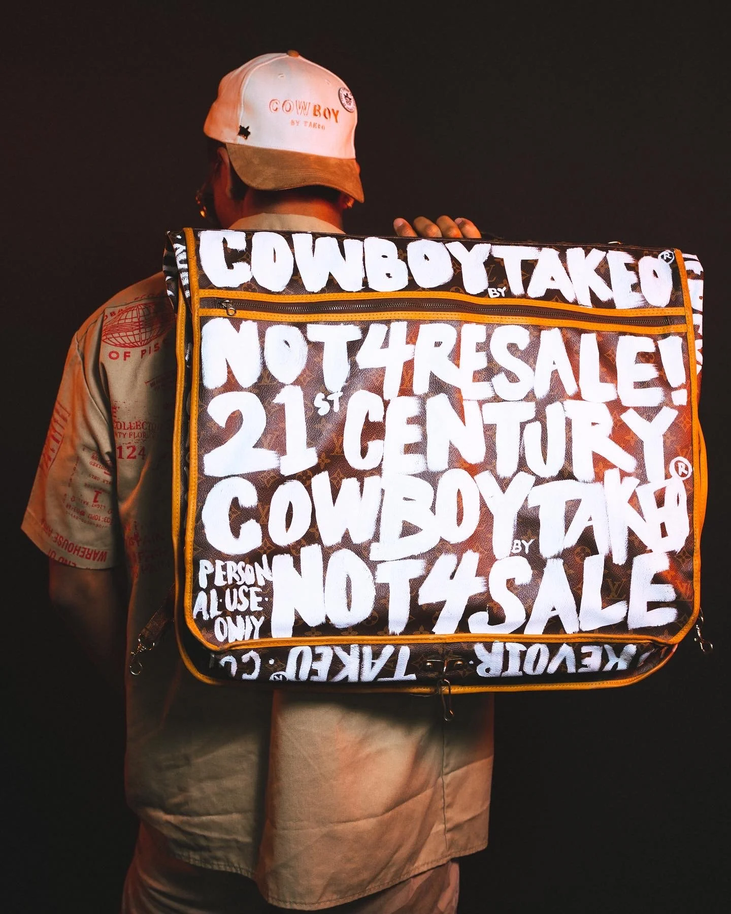 A person wearing a white cap with red and black text, and a beige shirt, is carrying a large black bag with white text that reads 'Cowboy Take Not 4 Sale! 21st Century Cowboy by Takeo' and additional smaller text, against a dark background.