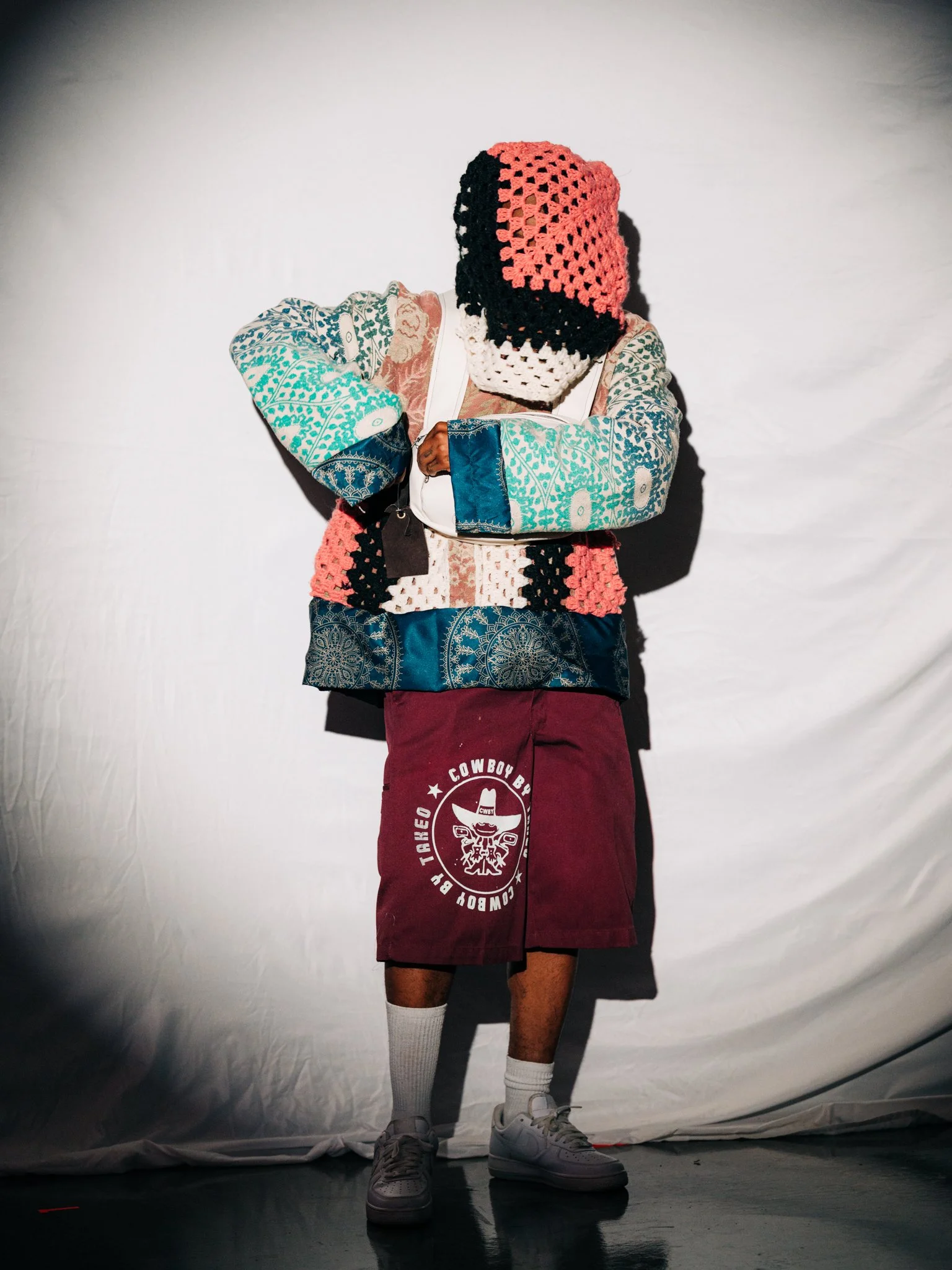 Person wearing a large, colorful patchwork jacket, maroon shorts with a cowboy logo, white socks, and sneakers, standing against a white backdrop, with arms crossed.