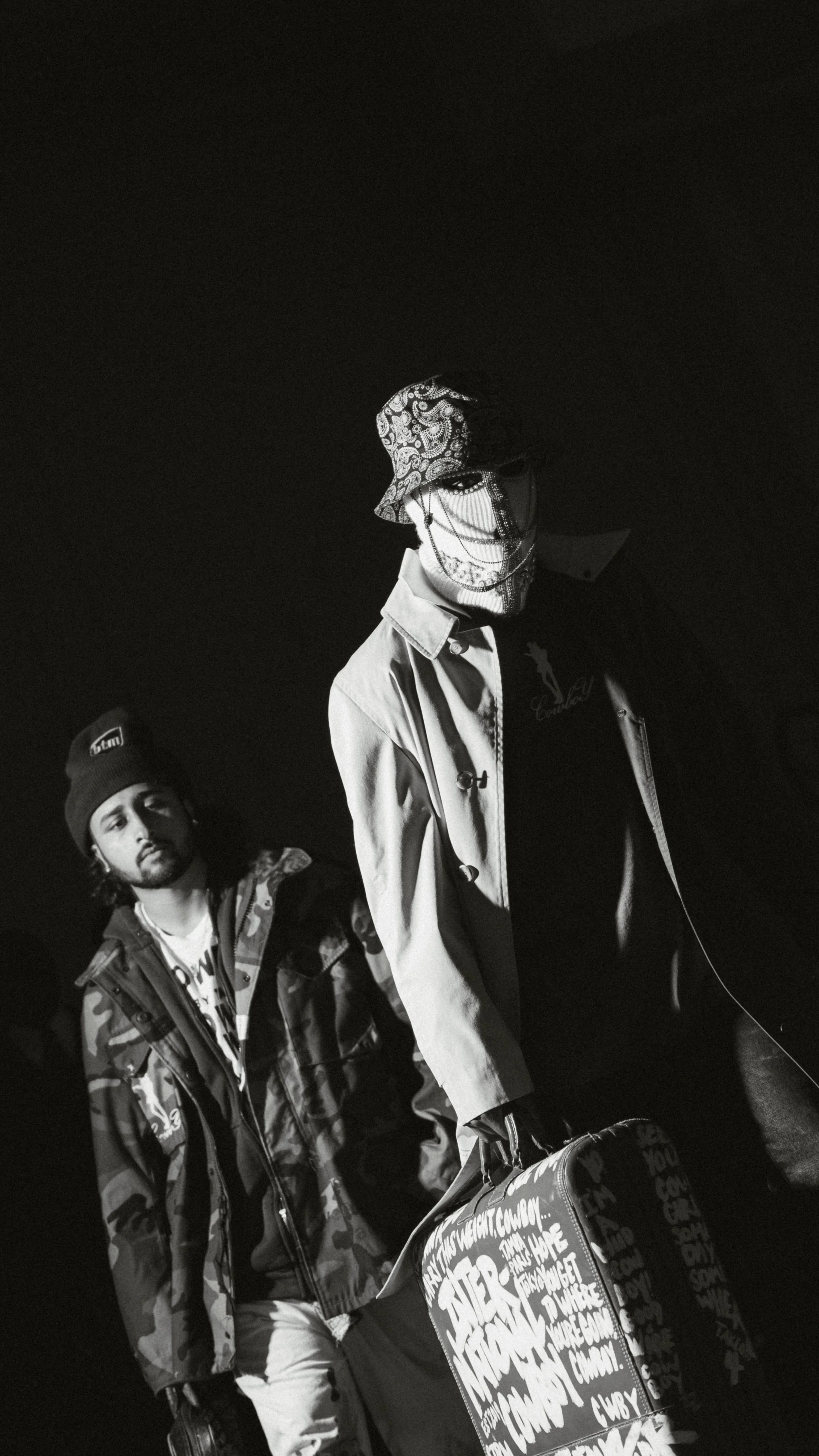 Two young men in streetwear standing in a dark setting, one holding a suitcase with graffiti-style text, both wearing jackets and accessories.