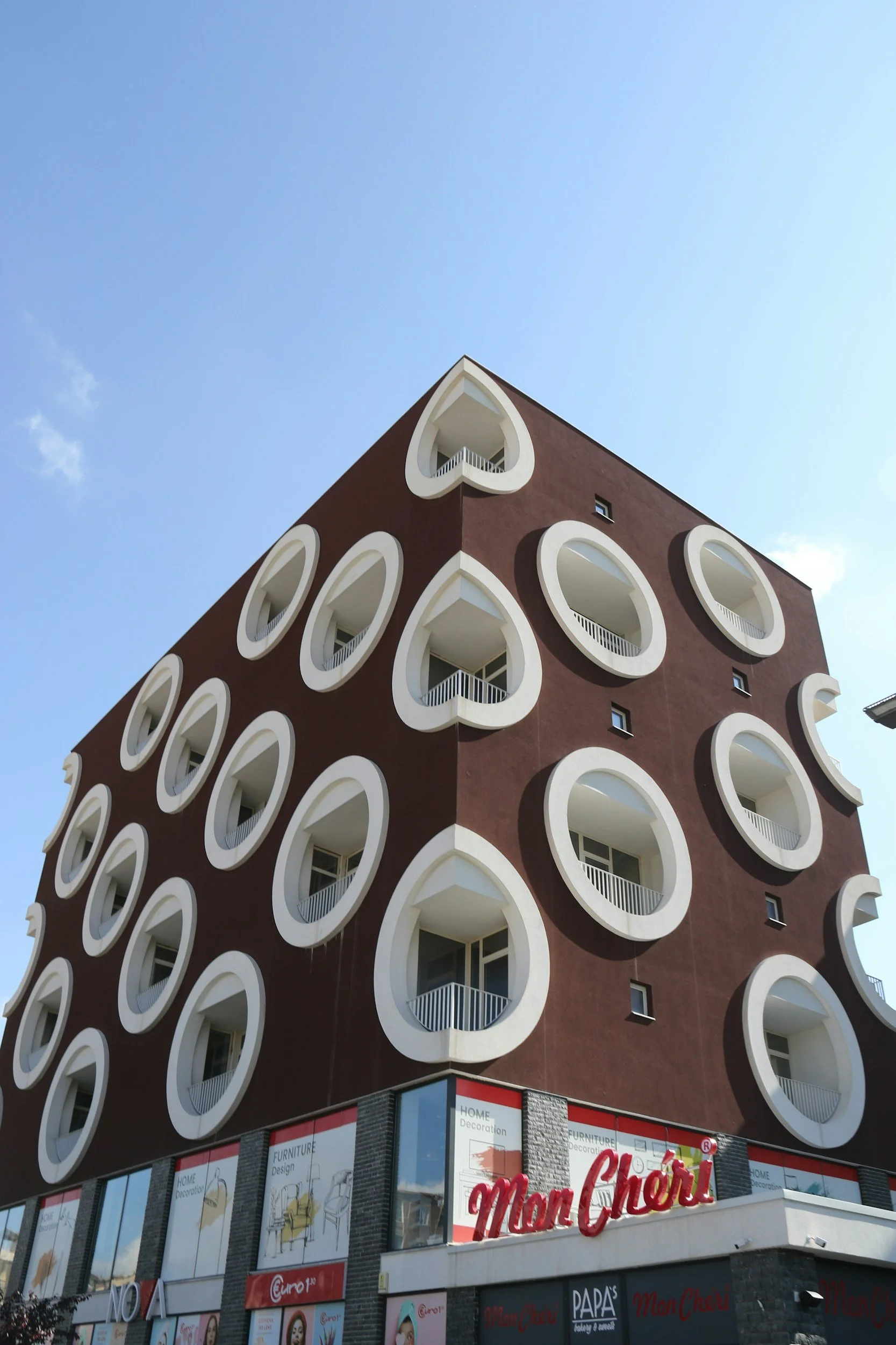 A five-story building with an unusual architectural design featuring multiple white, rounded window frames on a brown facade, with a retail store on the ground floor and a clear blue sky above.
