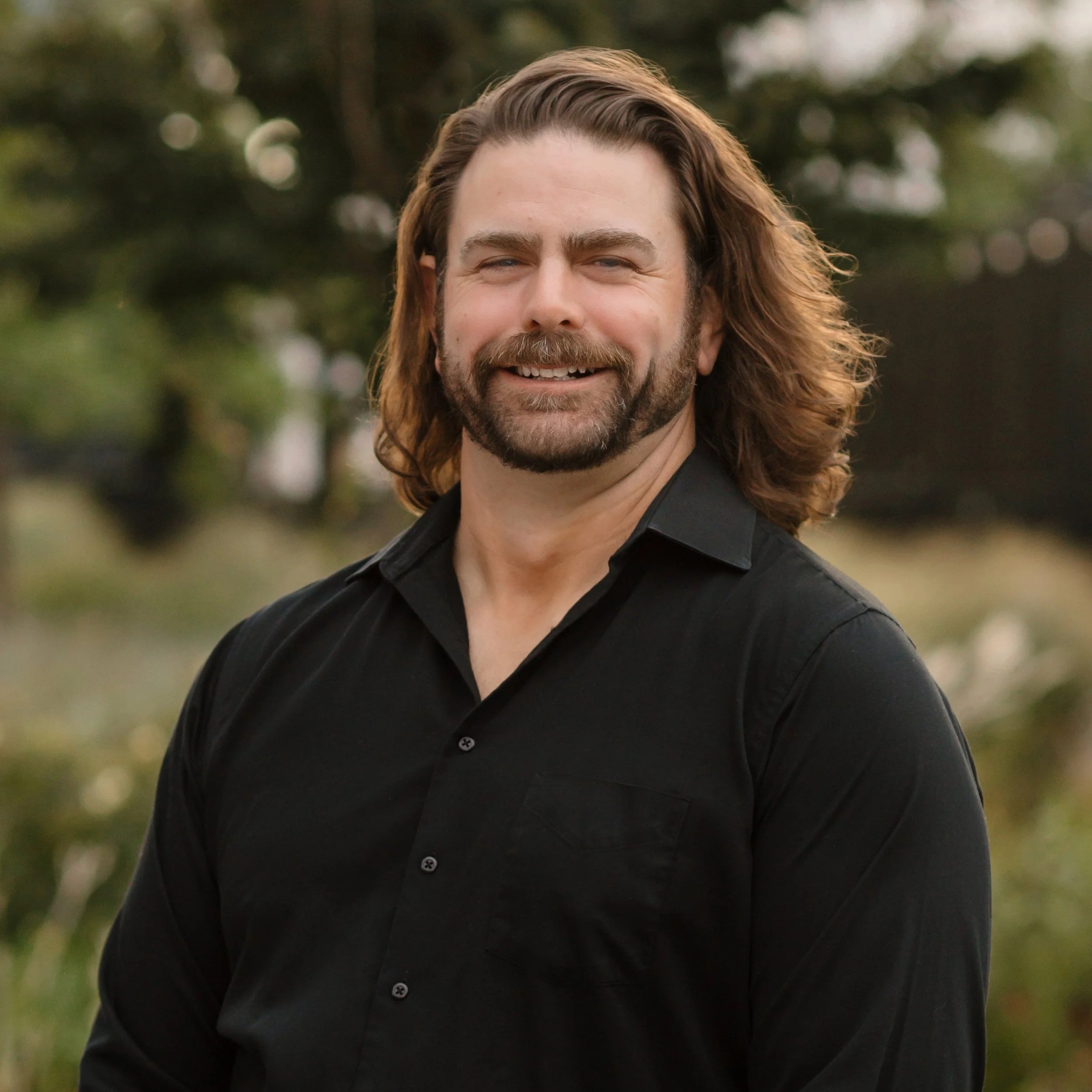 A man with long, wavy brown hair and a beard, wearing a black button-up shirt, standing outdoors with trees and greenery in the background.