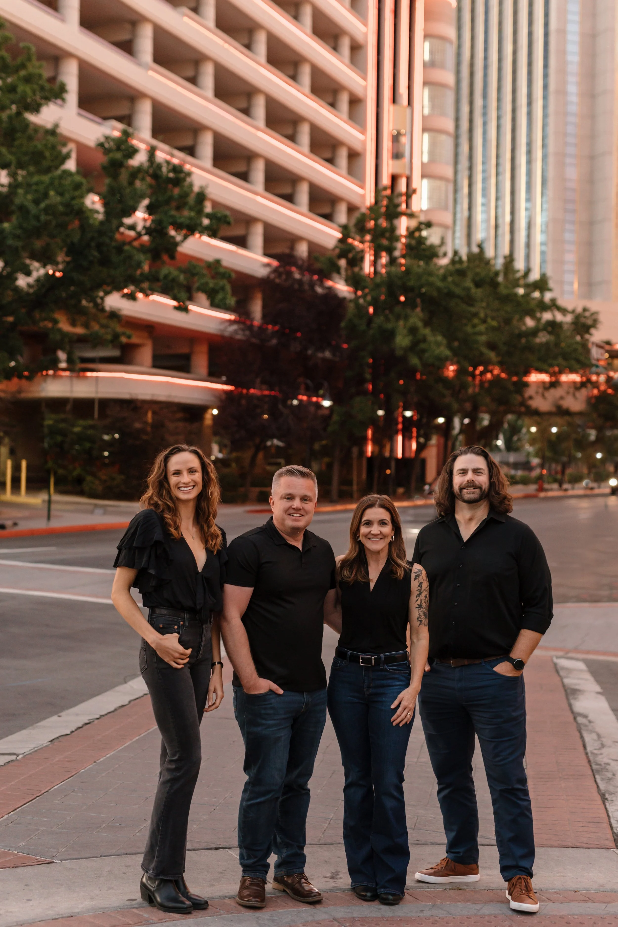 Four people standing on a city street at sunset, smiling at the camera with tall buildings and trees behind them.