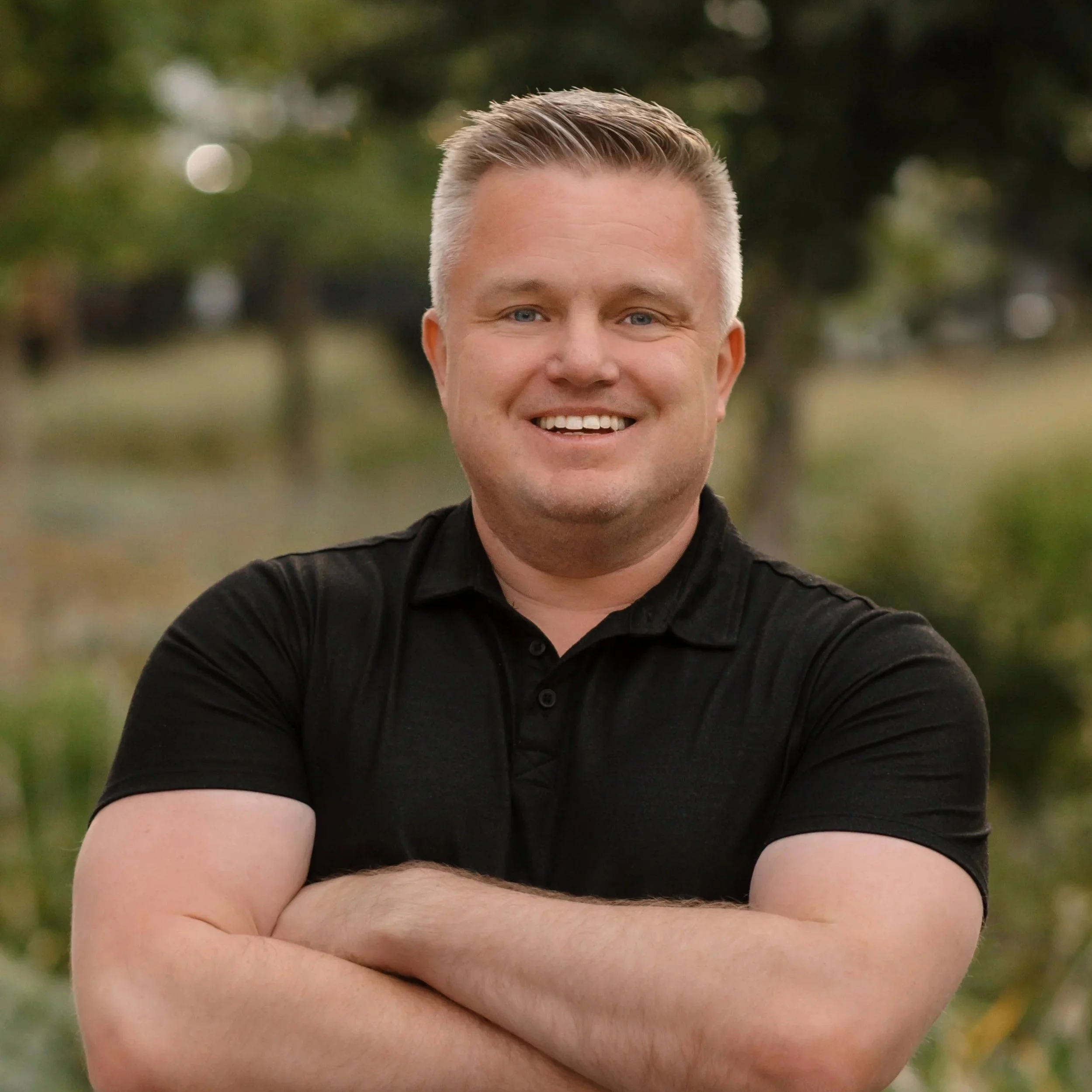 A smiling man with short, light-colored hair, wearing a black polo shirt, standing outdoors with trees and greenery in the background.