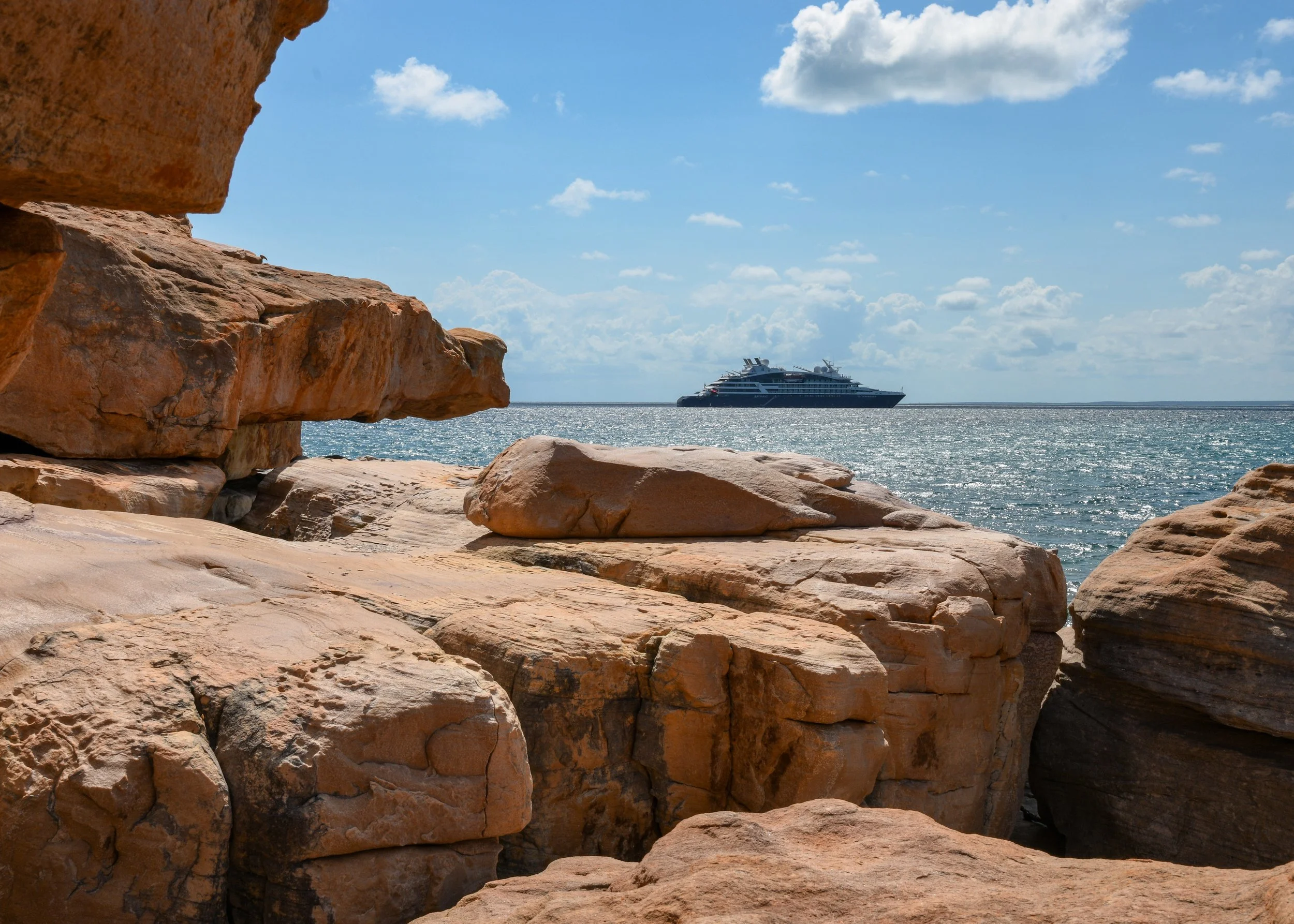 View of a large yacht on the ocean seen from between reddish-brown rocks on a sunny day with a blue sky and scattered clouds.