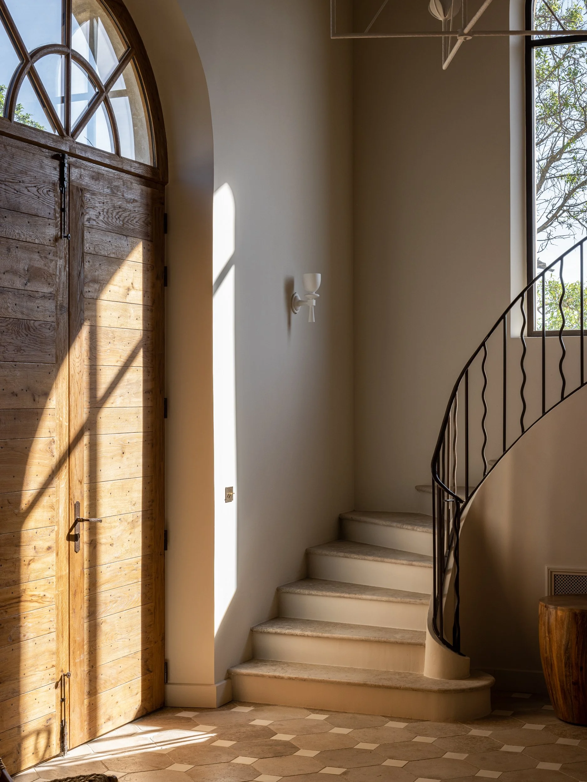 Sunlight streams through a large arched wooden front door into a hallway with tiled flooring, a spiral staircase with a black wrought iron railing, and a window with visible tree branches outside.