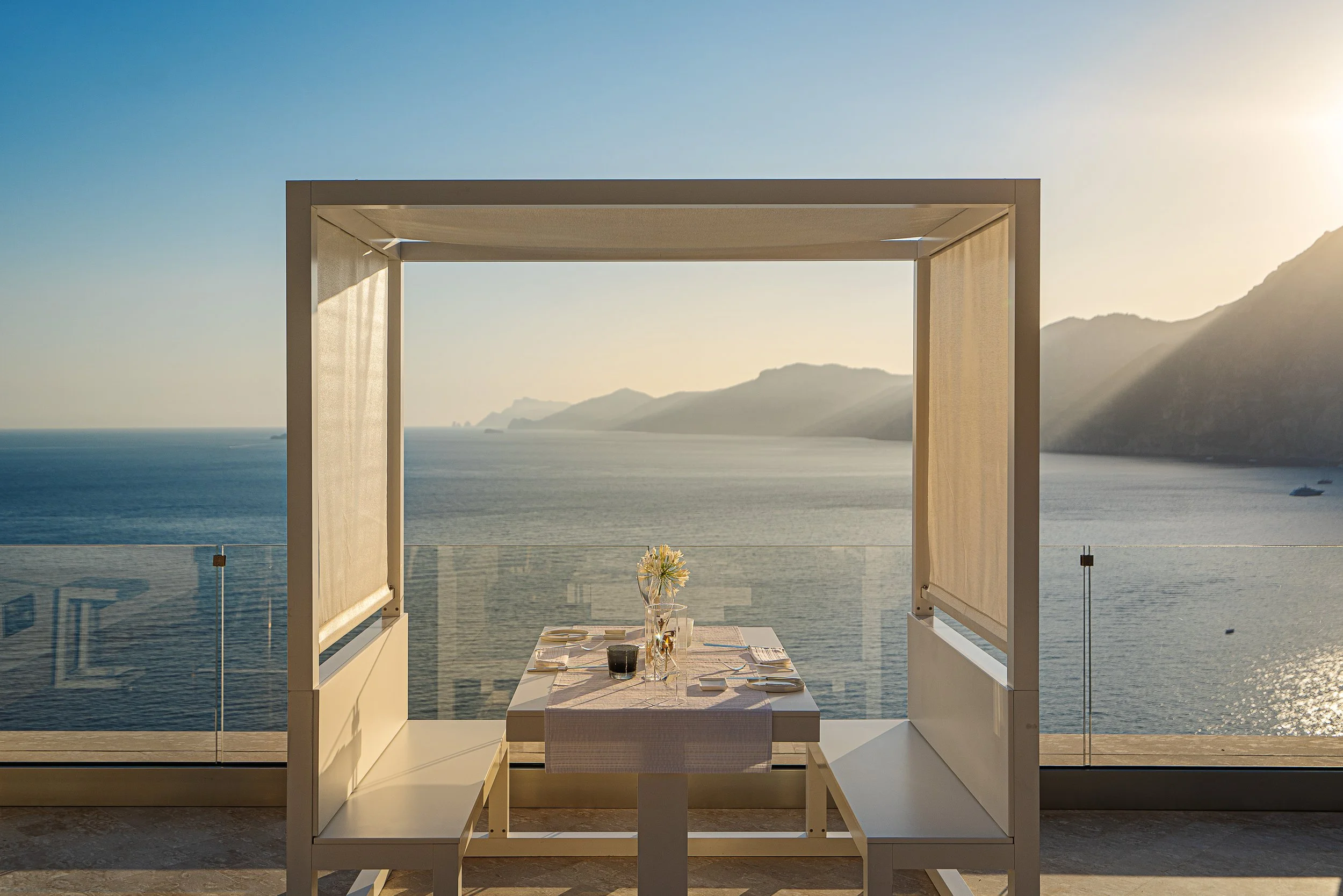 A table set for dining on a balcony overlooking a calm ocean with mountains in the distance during sunset.