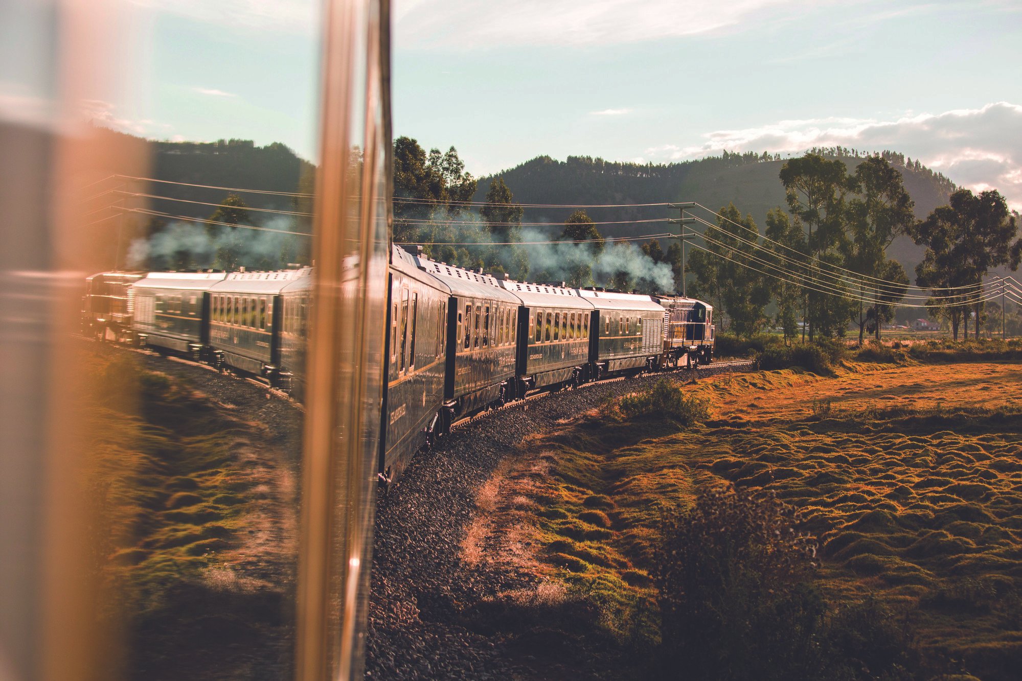 A train traveling through a scenic landscape with trees, hills, and power lines under a partly cloudy sky, reflected partially in a window.