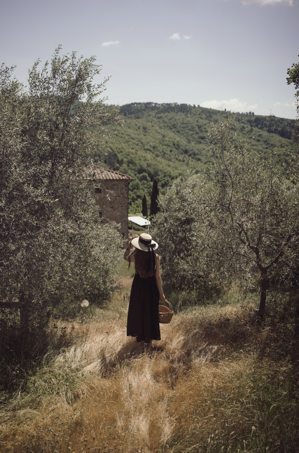 A woman wearing a black dress and a wide-brimmed straw hat walking through a sunny countryside path with olive trees, carrying a wicker basket, with green hills in the background.