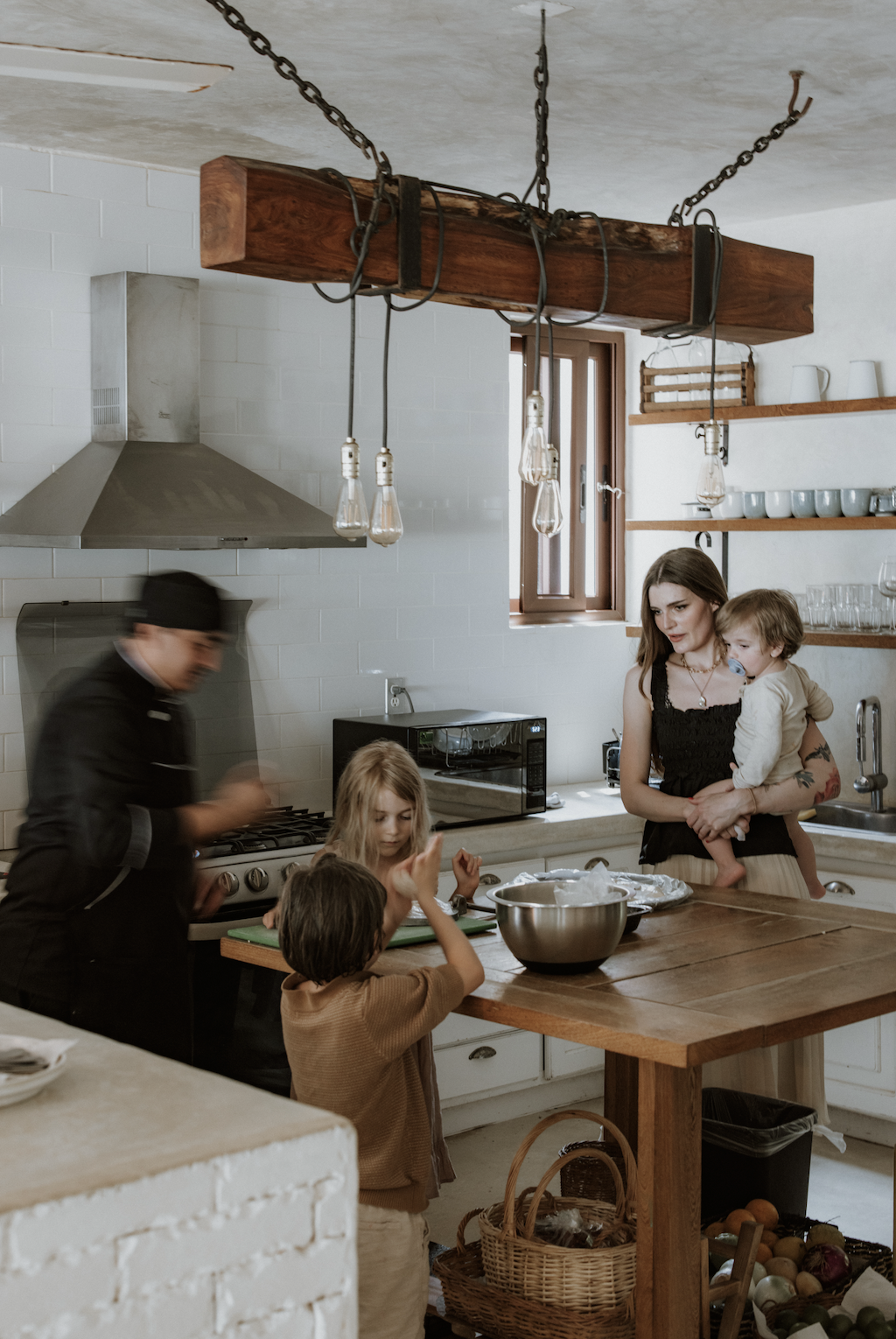 A family and a chef in a kitchen preparing food, with children around a table, and kitchen shelves with dishes in the background.