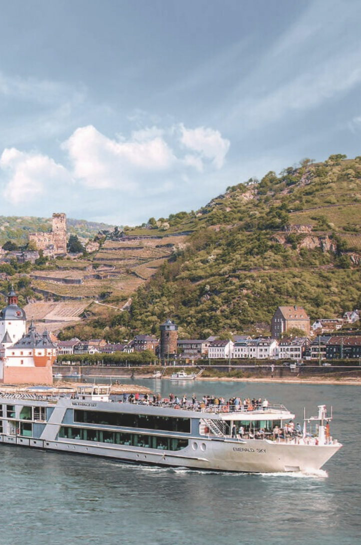A large boat named Emerald Sky sailing on the river with a town and green hillside in the background.