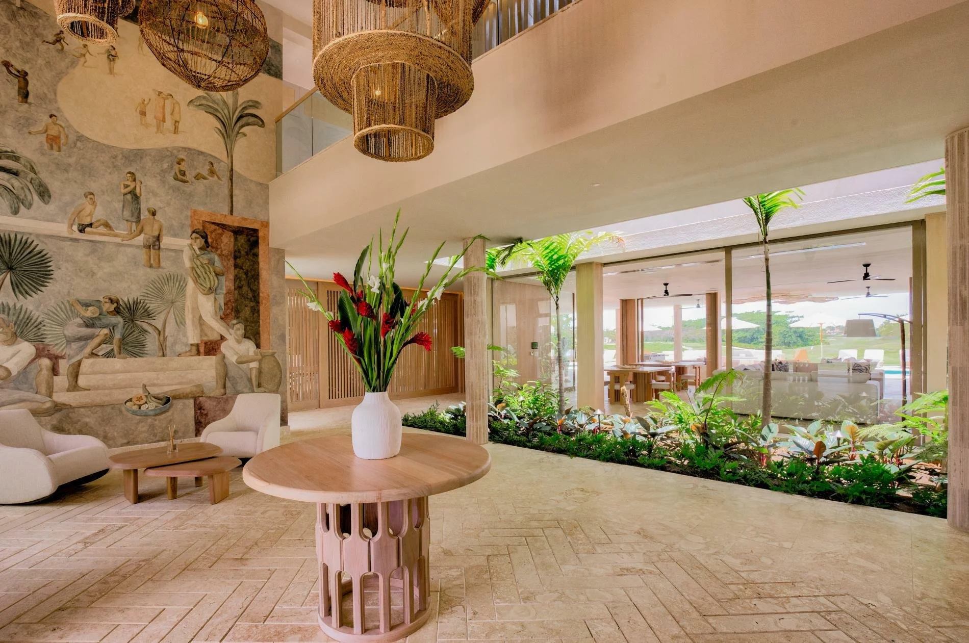 Modern hotel lobby with a large wall mural, a wooden table with a white vase of red and green flowers, white chairs, and an indoor garden with tropical plants and palm trees, overlooking an outdoor patio and pool area.