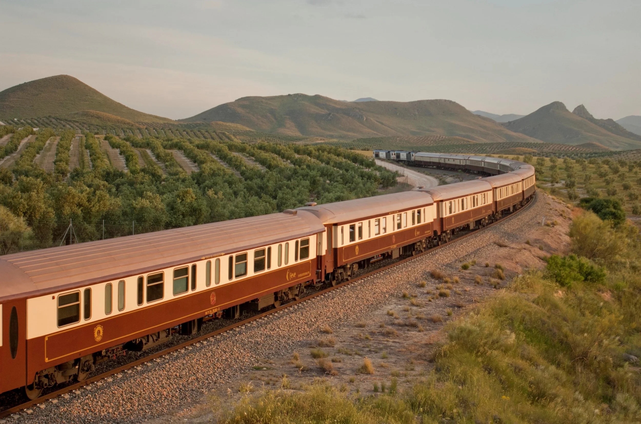 A vintage train with brown and cream cars traveling through a scenic landscape of rolling hills and green fields under a partly cloudy sky.