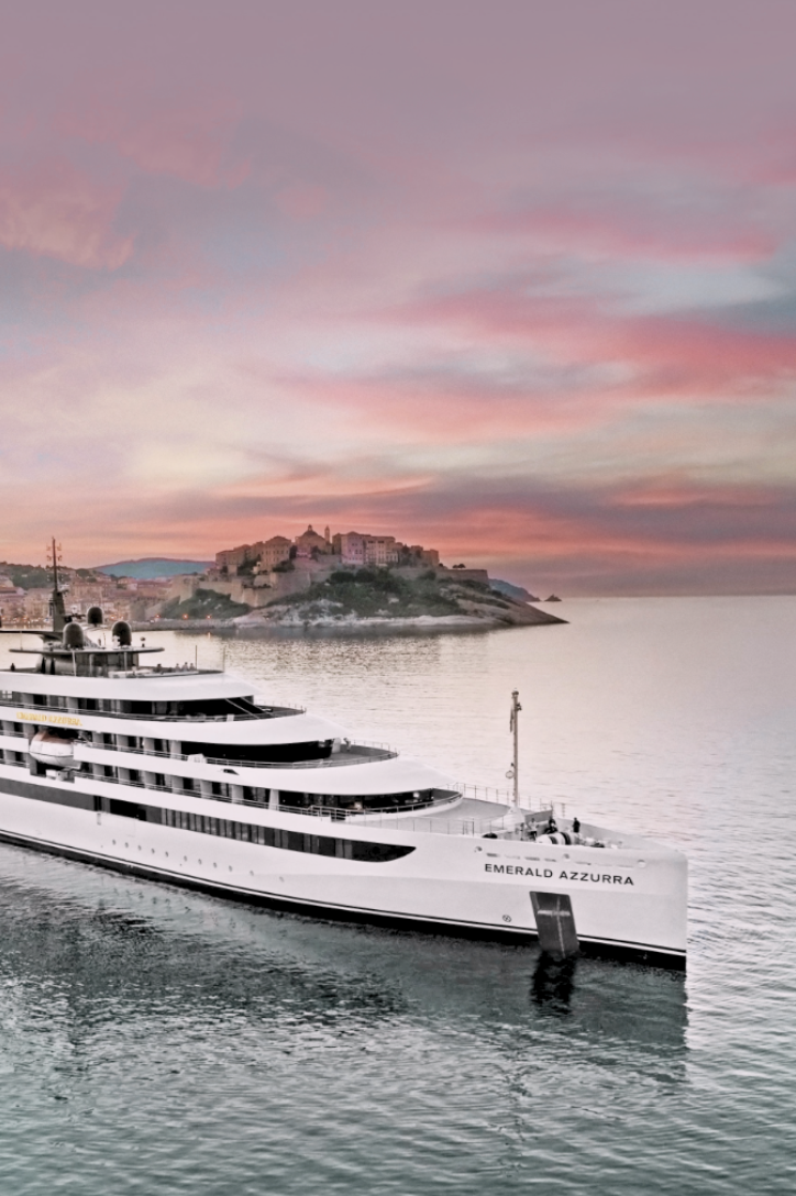 A large white yacht named Emerald Azzurra floating in a calm harbor with a hillside fortress and pink sunset sky in the background.