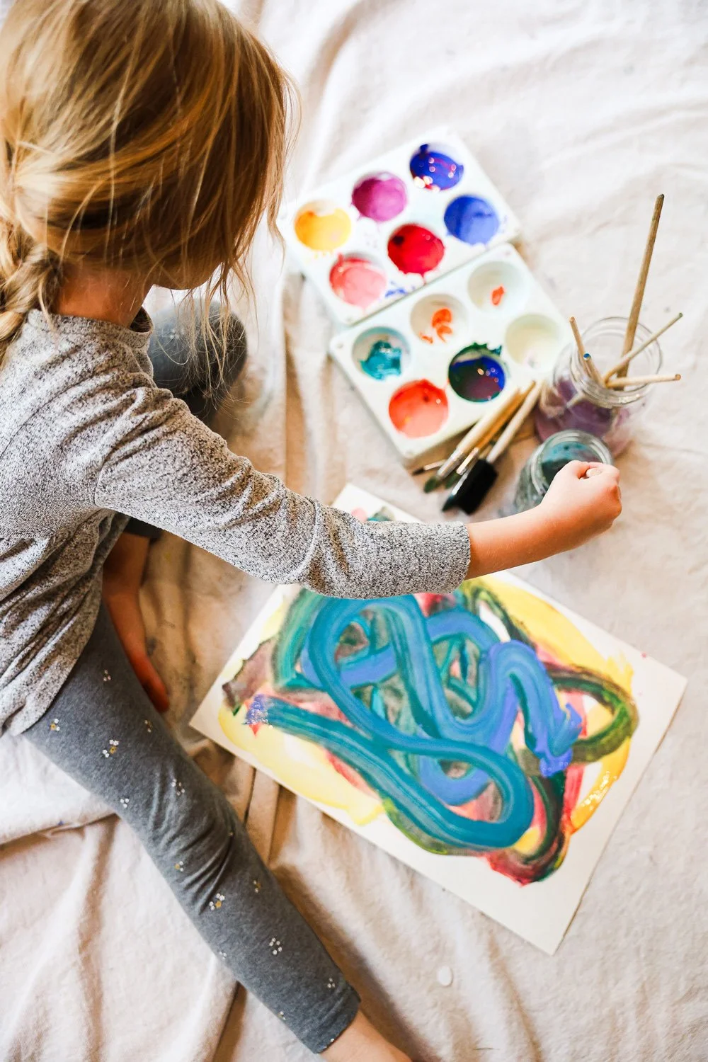 Child painting abstract art with blue, green, yellow, and red colors on paper, surrounded by watercolor paints and brushes.