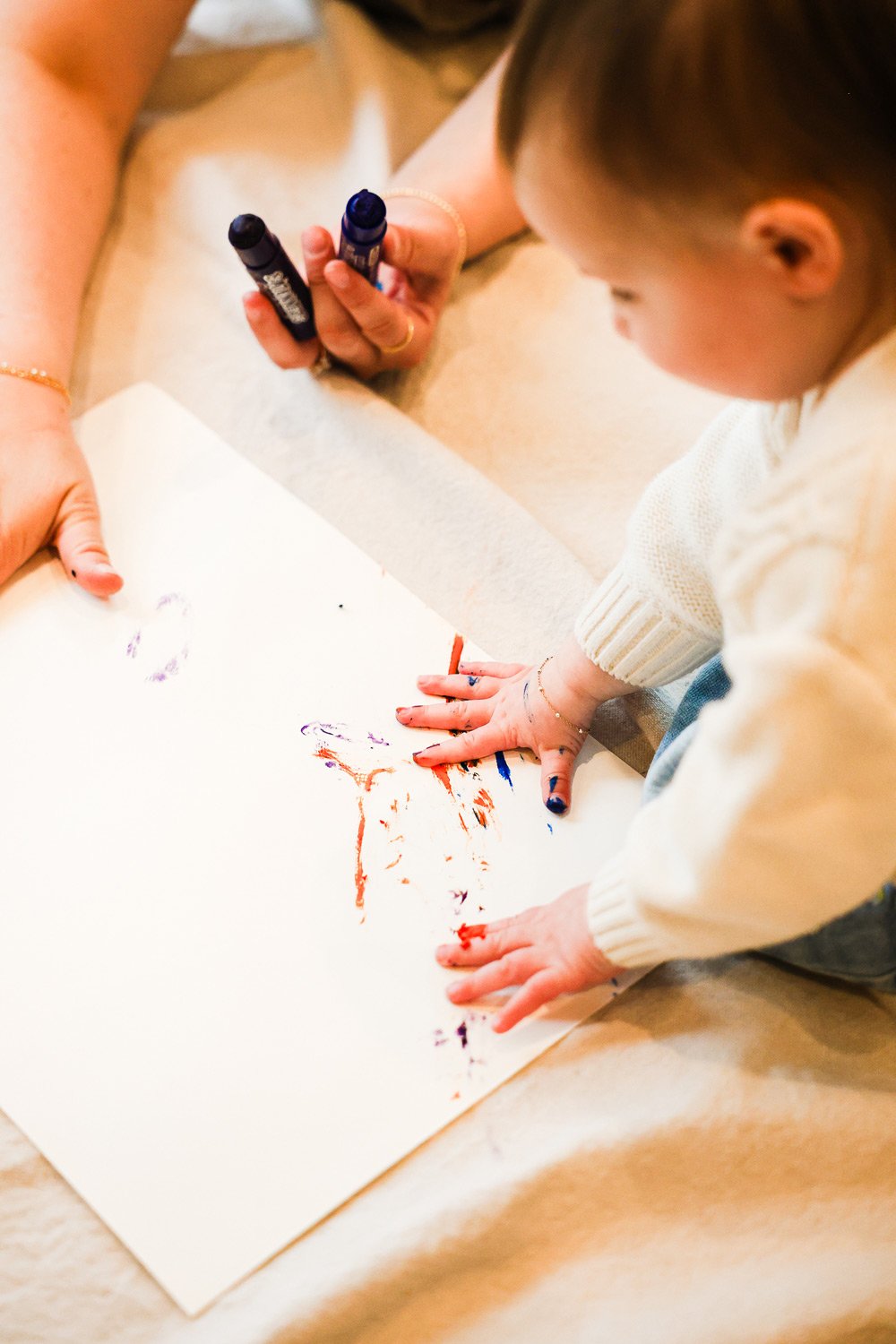 A young girl with paint on her hands and fingers, leaning over a large sheet of paper on the floor, coloring with markers during an art activity.