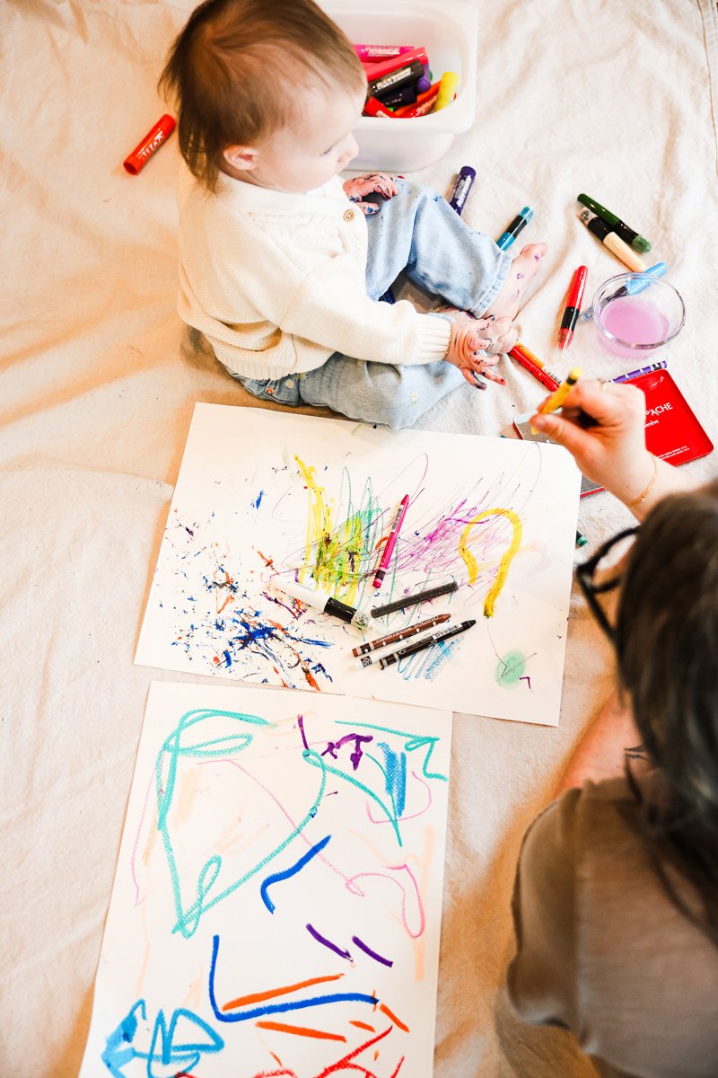 Child sitting on a beige blanket creating colorful abstract drawings with crayons, surrounded by art supplies.