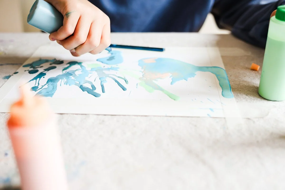 Person creating a watercolor painting on paper with blue and green paints, with bottles of paint and a paintbrush on a table.