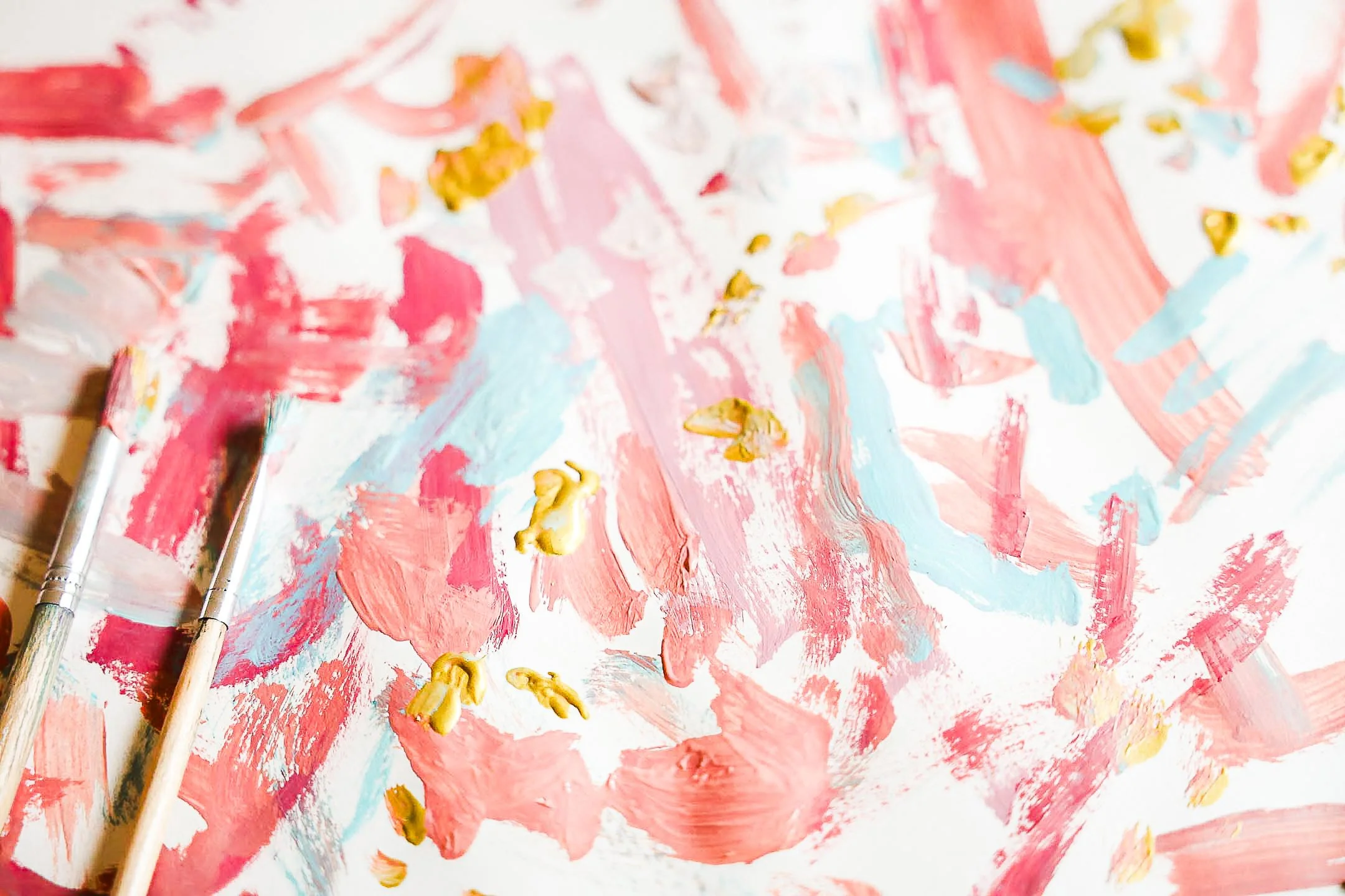 Abstract painting with pink, blue, white, and gold paint, alongside two paintbrushes.
