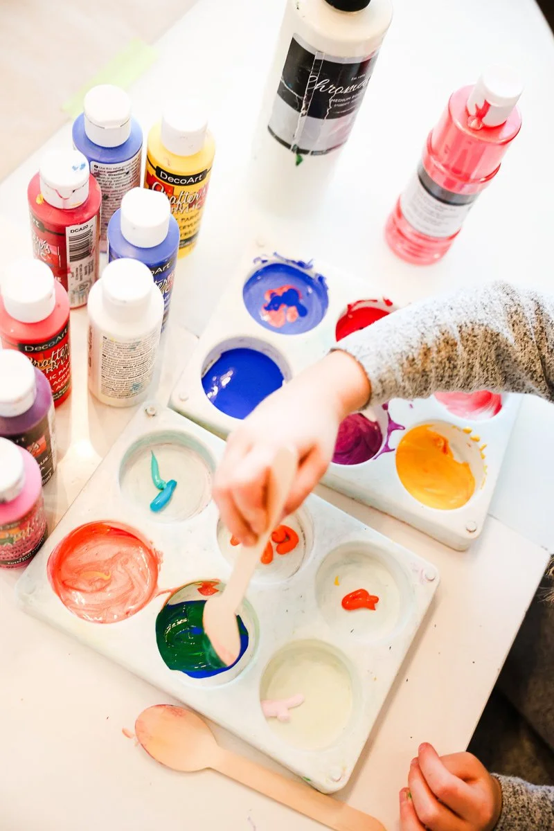 Child mixing colorful paints in a palette with bottles of paint surrounding the workspace.