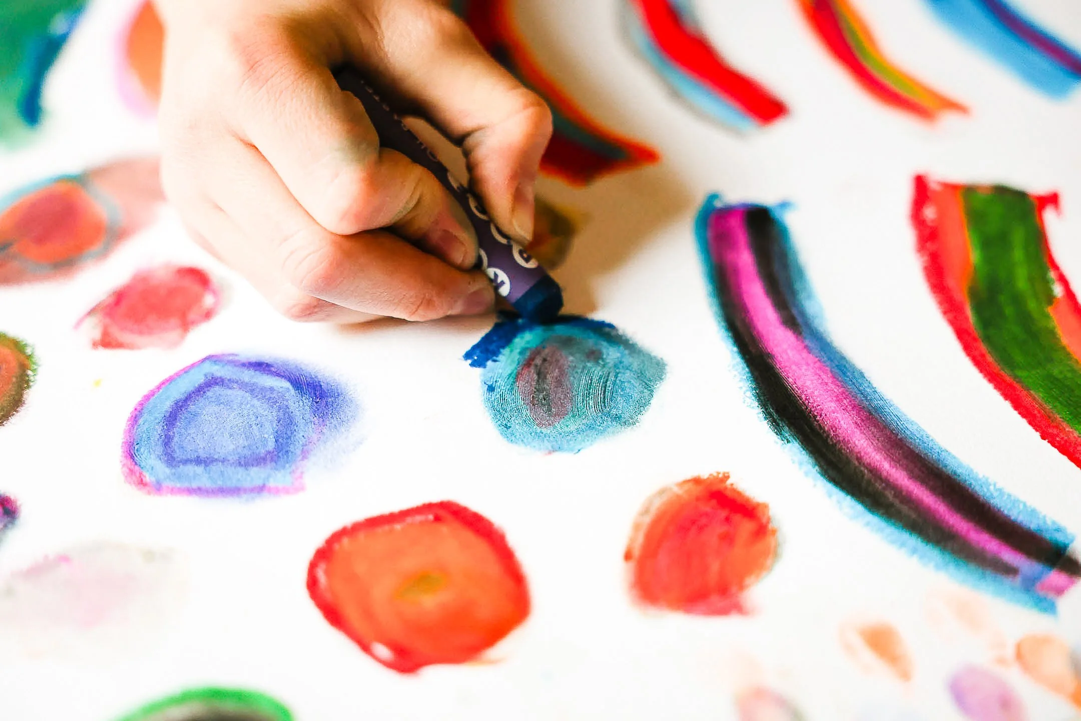 A person's hand holding a black crayon, drawing colorful watercolor shapes and lines on a white sheet of paper, with various vibrant colors and patterns.