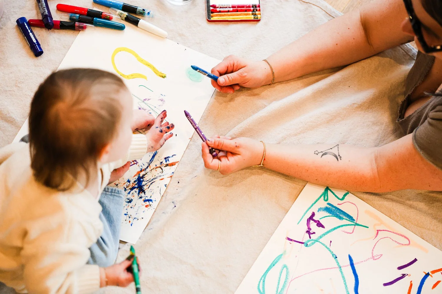 A child and an adult sitting at a table, drawing and coloring on large sheets of paper with markers. The table is scattered with colorful markers, and their hands are engaged in creative art activities.
