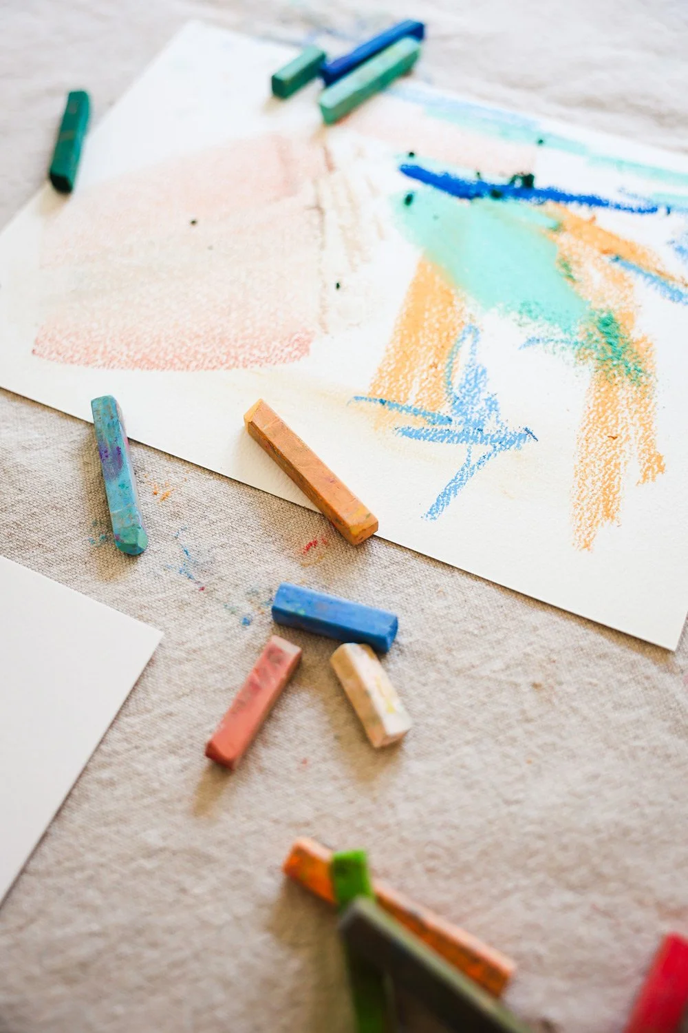 A sheet of paper with colorful pastel chalk drawings and several pastel chalks scattered around on a light fabric surface.