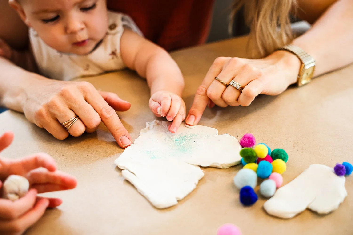 Two adults and a young child decorating shaped sugar cookies with colorful sprinkles and pom-poms on a brown table.