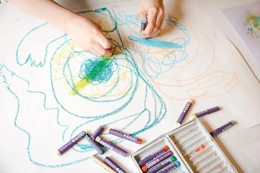 Children's hands drawing colorful scribbles with oil pastels on a large piece of white paper.