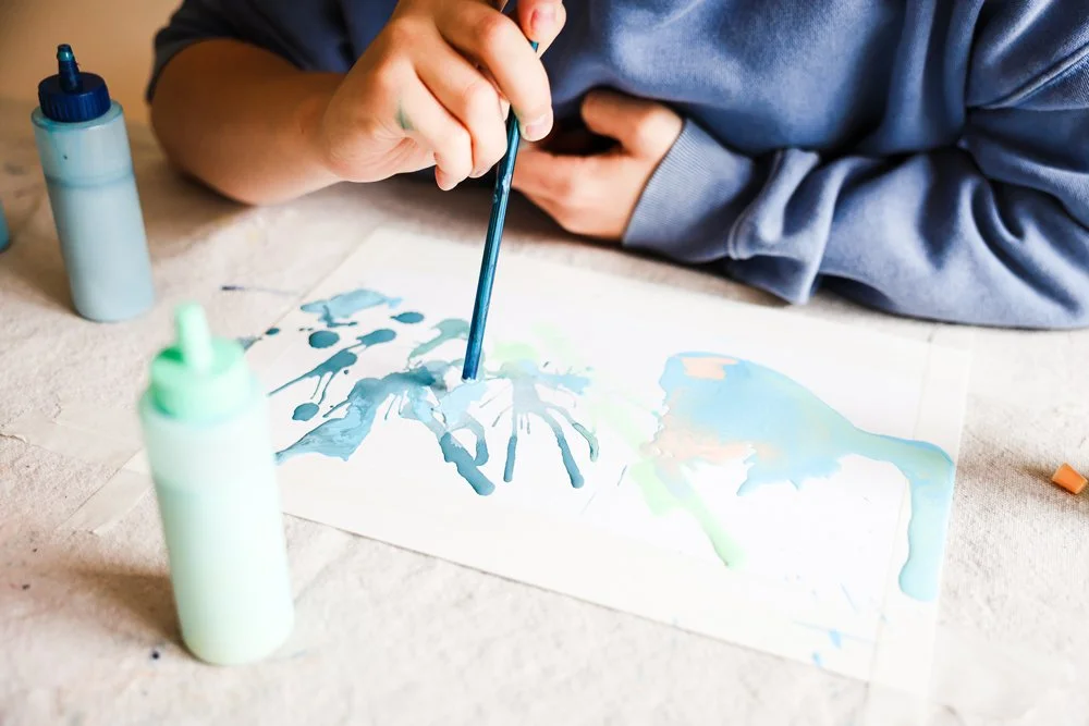 Child creating watercolor art with bottles of paint on paper.
